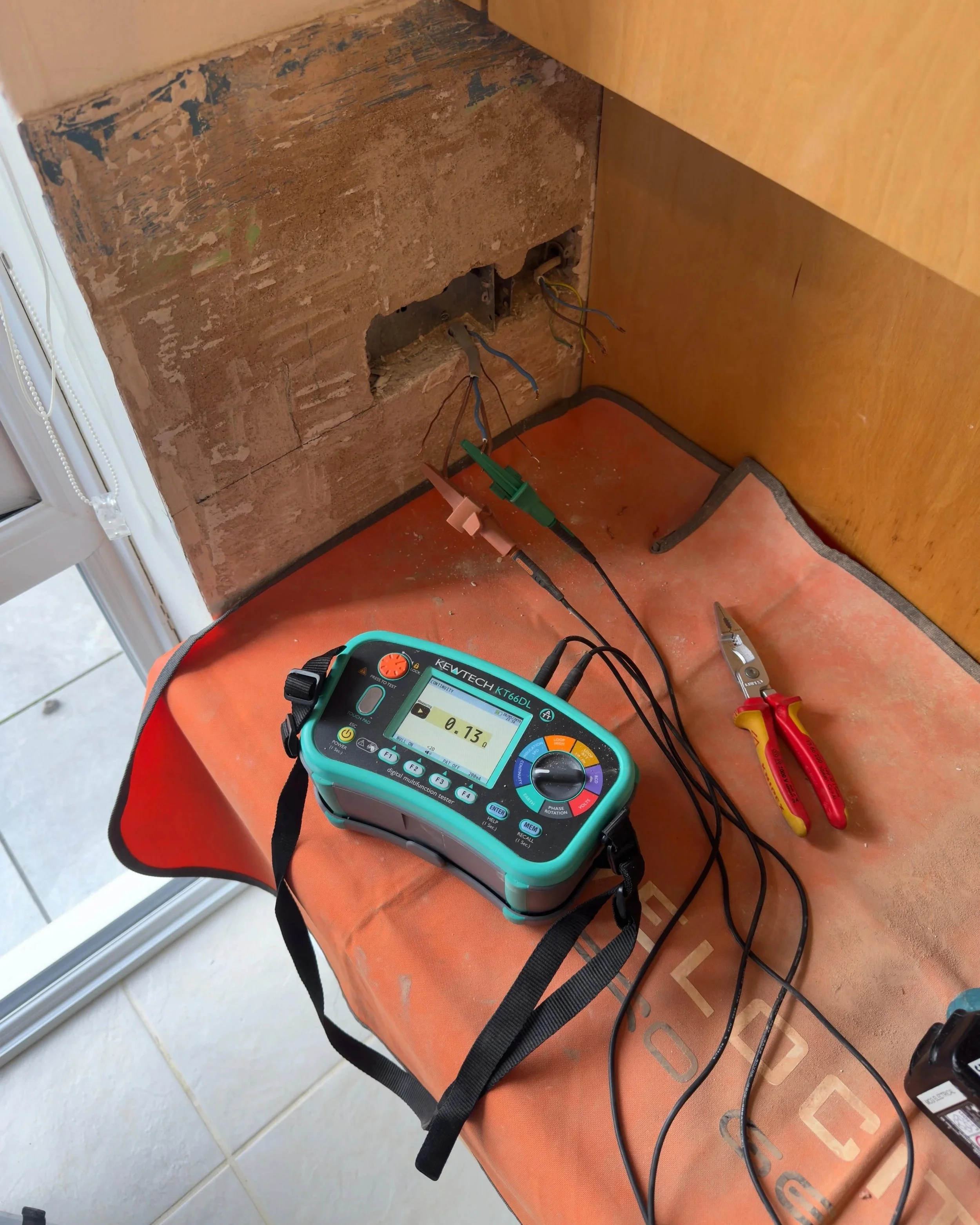 Electrical wiring being tested with a multimeter connected to a wall outlet. There are wires, pliers, and testing equipment on a work surface.