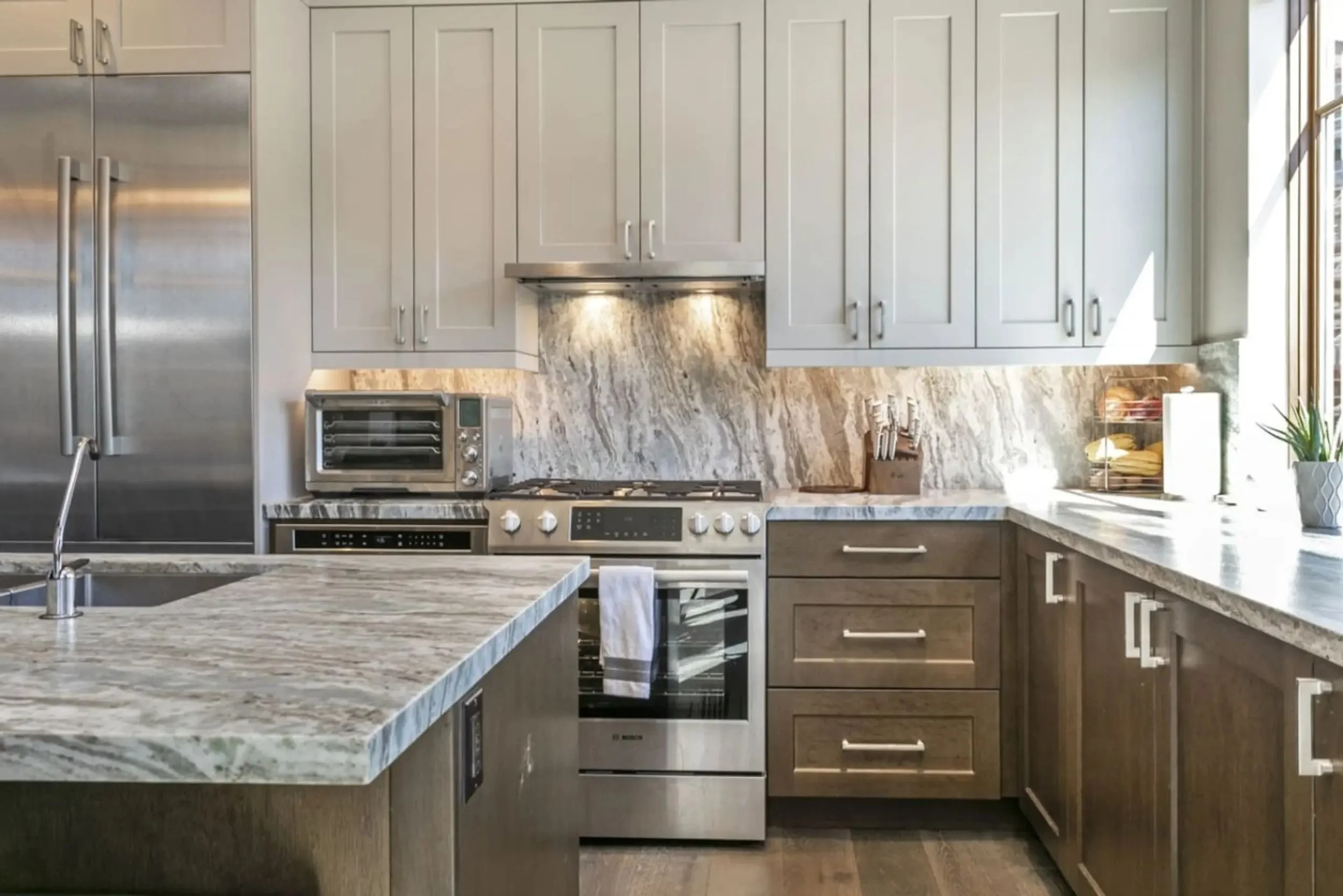 Modern kitchen with a granite countertop island, stainless steel refrigerator, oven, microwave, and a window with sunlight. There are upper and lower cabinets in gray and wood finish.