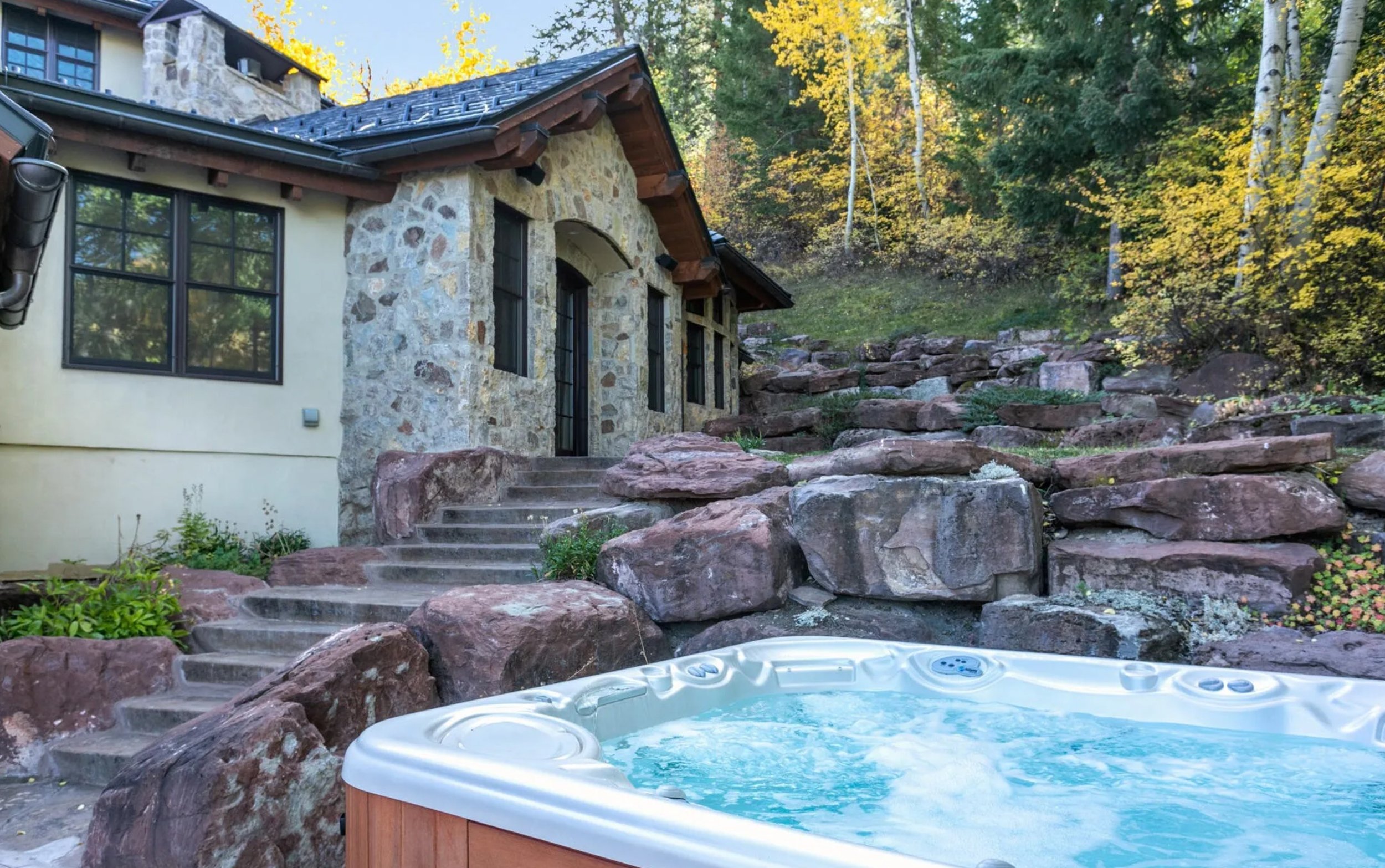 Stone and stucco house with steps leading up to the door, surrounded by rocks and landscaping, with a hot tub in the foreground, and trees with fall foliage in the background.
