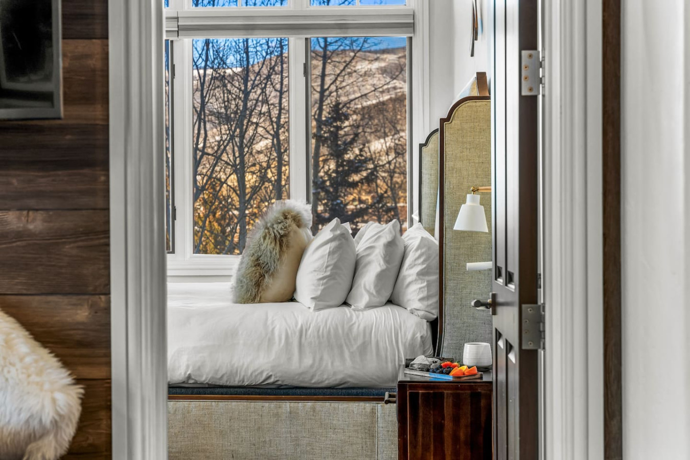 A cozy bedroom with a window view of trees and mountains during sunset, featuring a fluffy dog lying on a bed with white pillows.