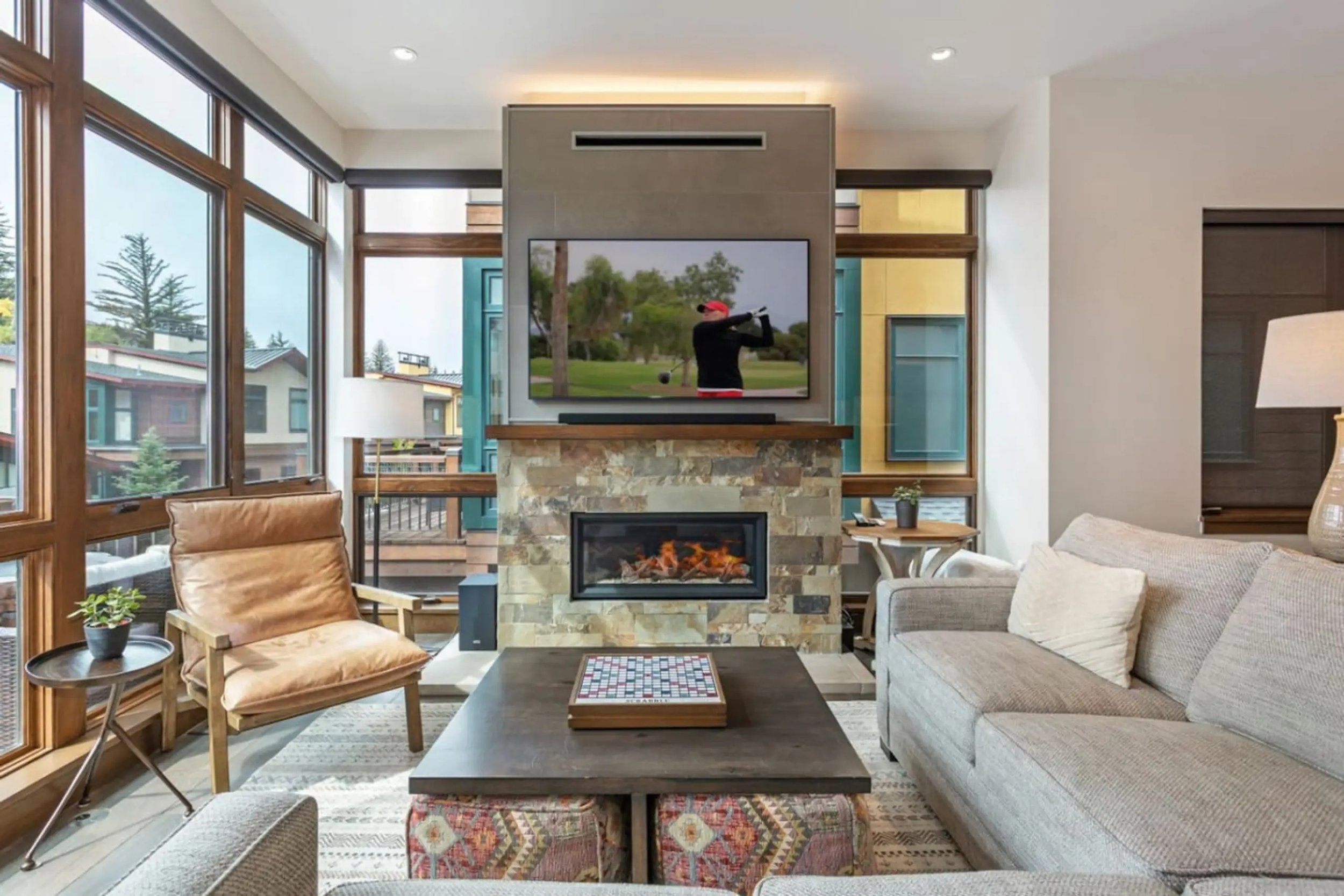 Living room with large windows, beige couch, wooden armchair, gas fireplace, wall-mounted TV showing a golfer, side tables with lamps, and decorative pillows.