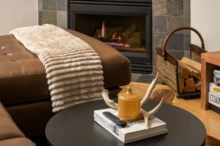 Living room with a fireplace, a brown leather ottoman with a cream-colored throw blanket, a round black coffee table with decorative items including a yellow teapot-shaped container and an antler-shaped object, a basket of firewood, and shelves with board games.