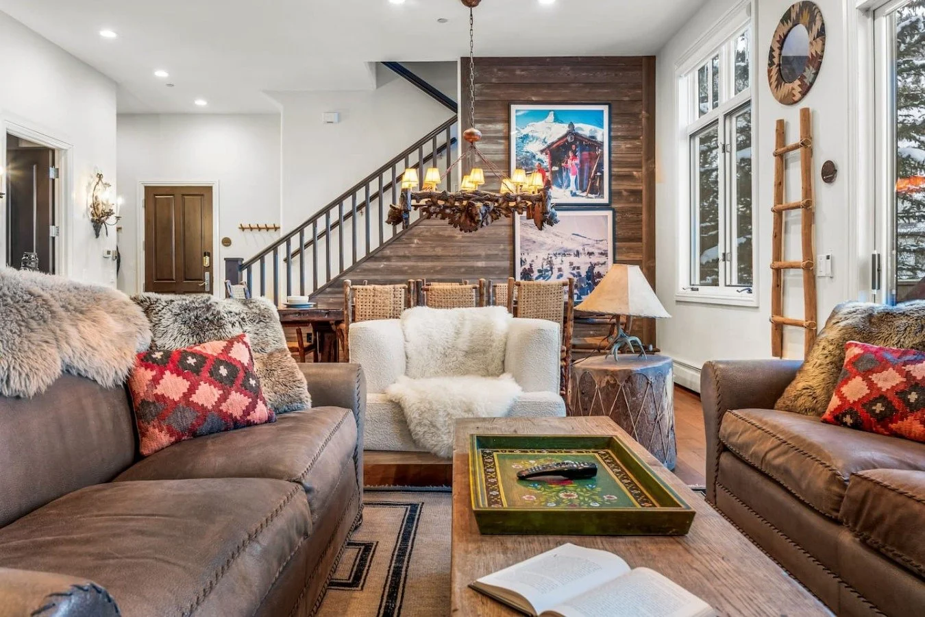 A cozy living room with two brown leather sofas, a white armchair with a fur blanket, and colorful throw pillows. There's a wooden coffee table with a game board, a book, and a remote. The space features wooden accents, large windows, and a staircase with a wooden railing leading upstairs. Wall art and a ladder decorate the room, creating a warm, rustic atmosphere.