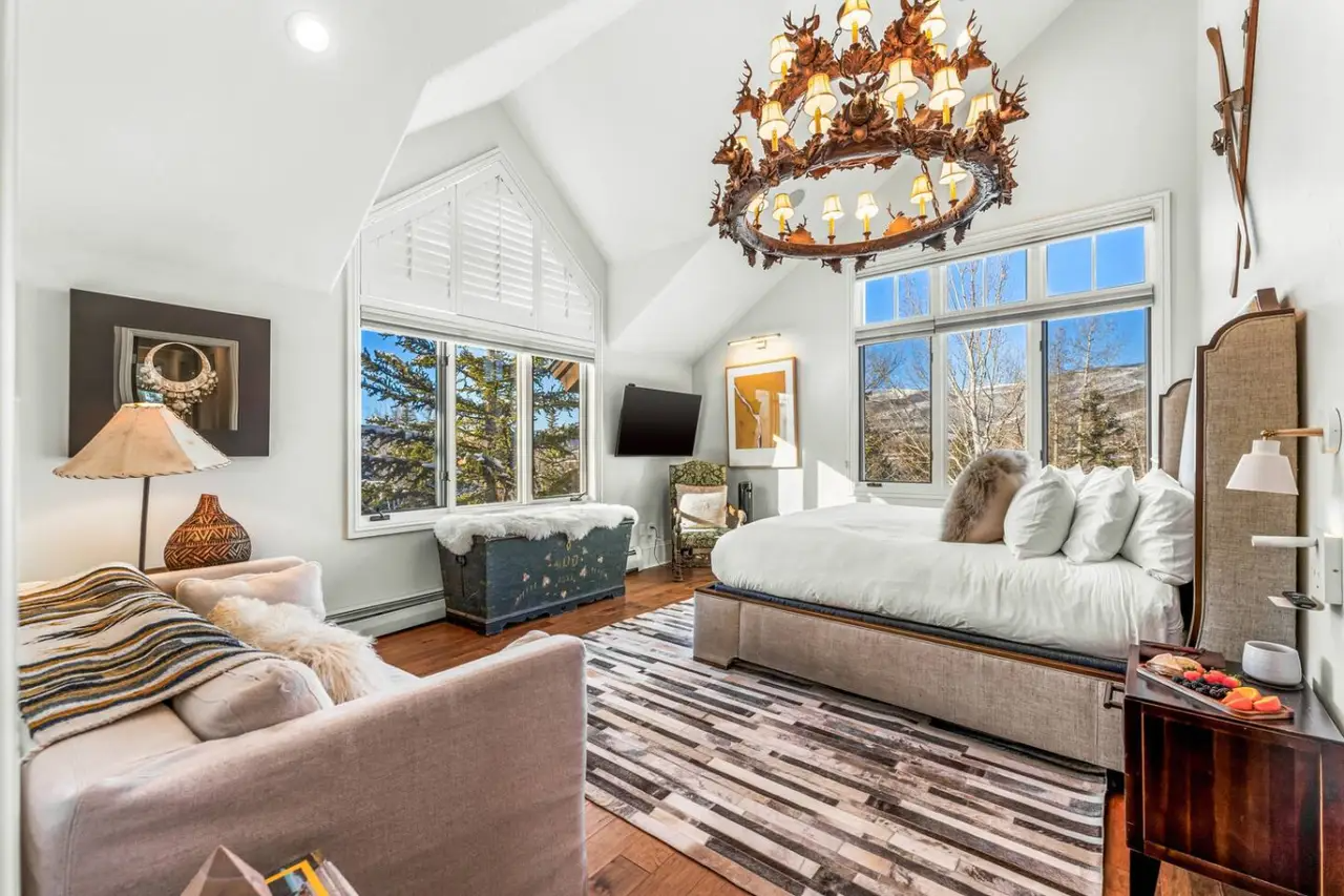 A bright bedroom with large windows, a white bed with pillows, a small sofa with throw blankets, a chandelier, a wooden side table, and a flat-screen TV mounted on the wall.