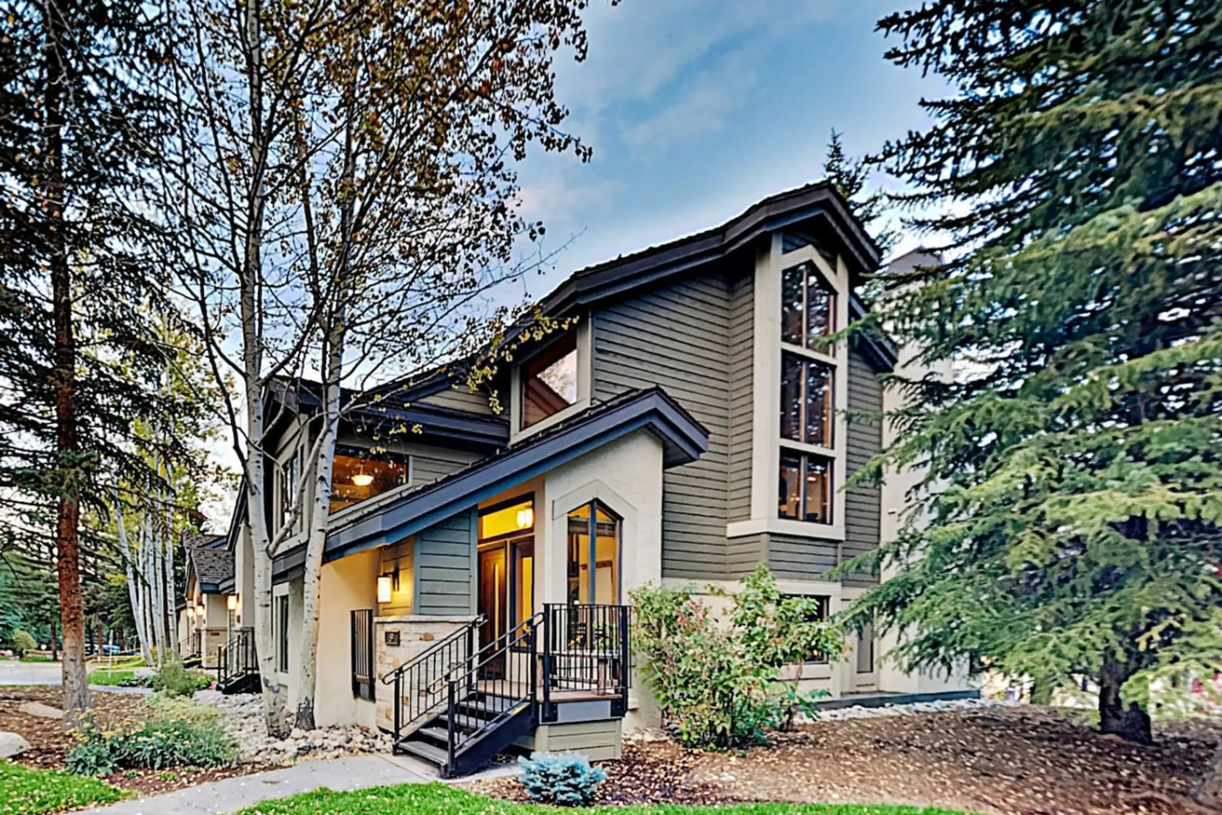 A modern multi-story house with gray and white exterior, surrounded by trees and greenery, with a small front porch and black railing, during daytime with a partly cloudy sky.