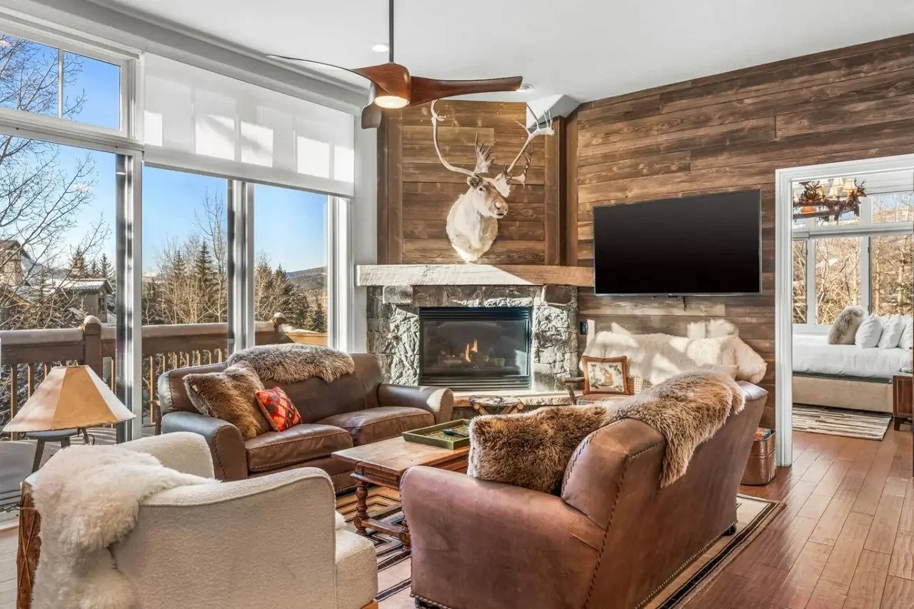 Cozy living room with large windows, stone fireplace, mounted deer head, leather and fabric sofas with fur throws, wooden coffee table, ceiling fan, and a flat-screen TV, overlooking a winter landscape outside.