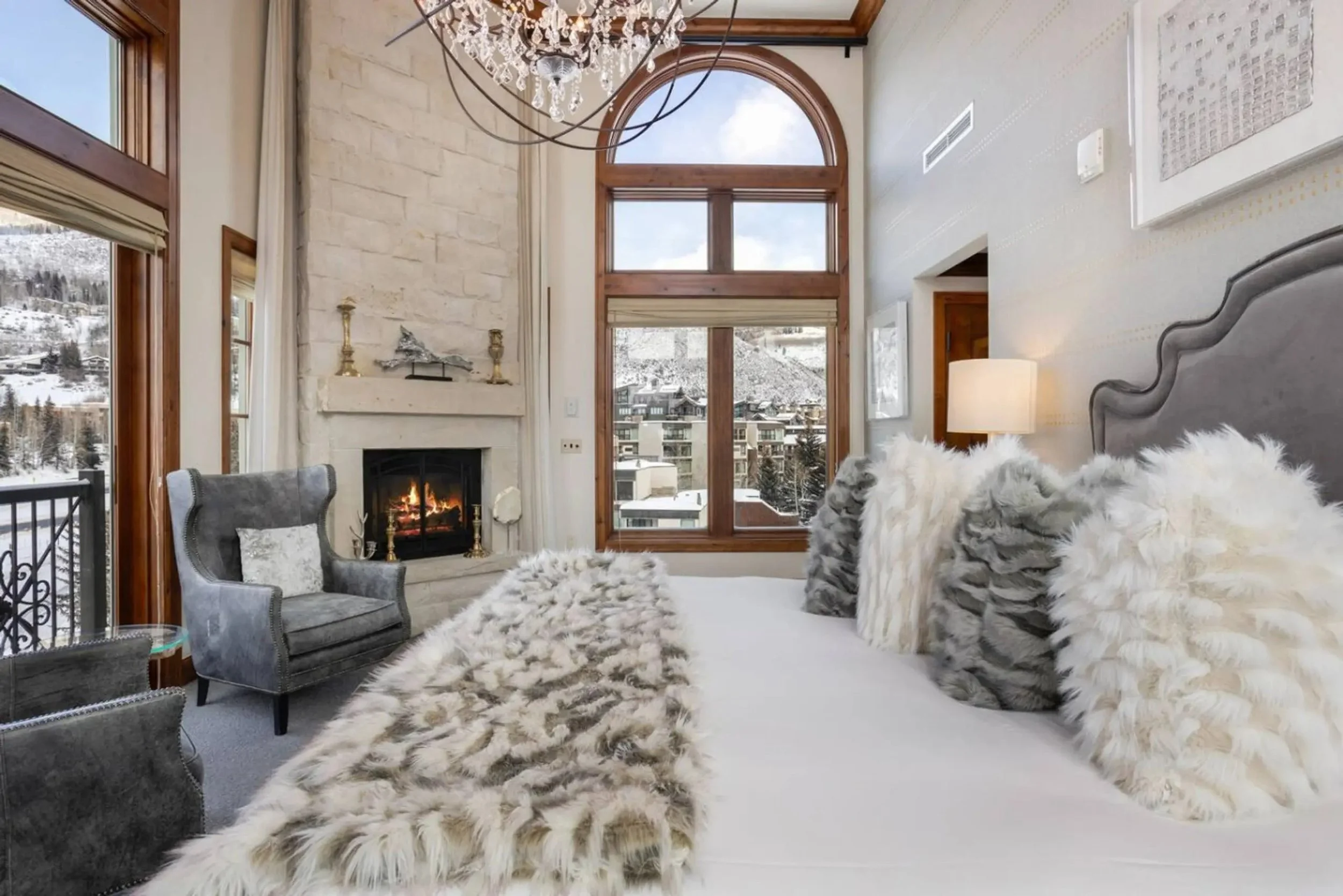 A cozy bedroom with large wooden framed windows showing a snowy outdoor landscape, a stone fireplace with a fire burning, and plush pillows and blankets in white and gray tones.