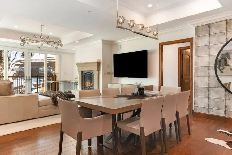 Modern dining room and living space with beige chairs around a wooden dining table, flat-screen TV on the wall, fireplace, large window with a snowy outdoor view, and contemporary lighting fixtures.