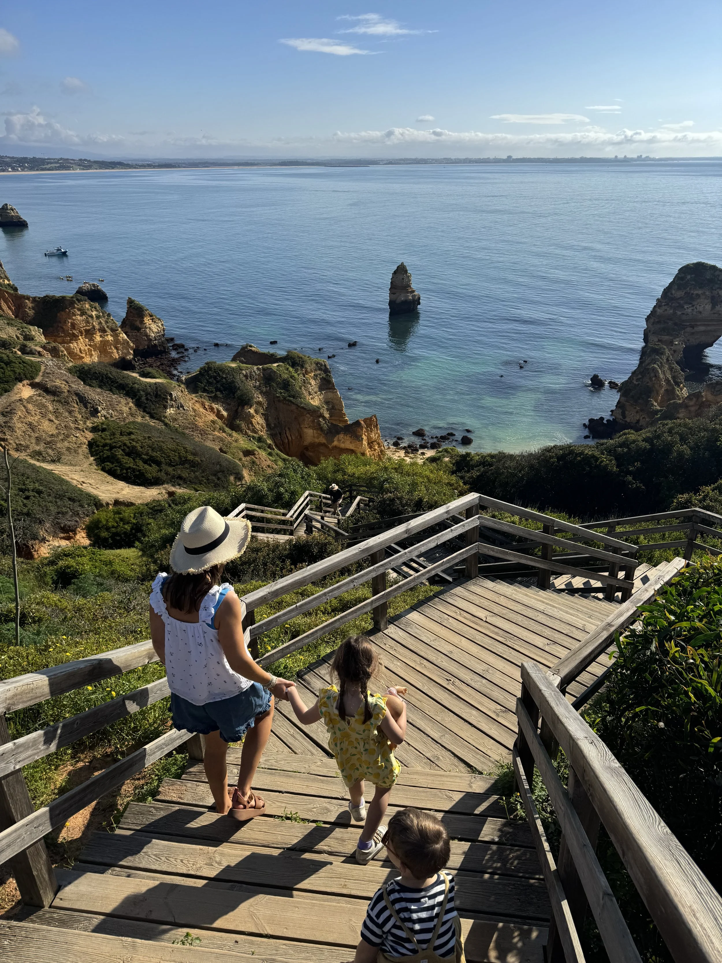 A woman and two children walking down wooden stairs towards the ocean at a scenic coastal overlook.