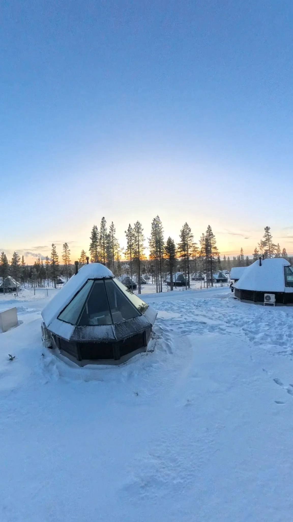 Snow-covered igloo and small cabins in a snowy landscape with a forest and sunset sky in the background.