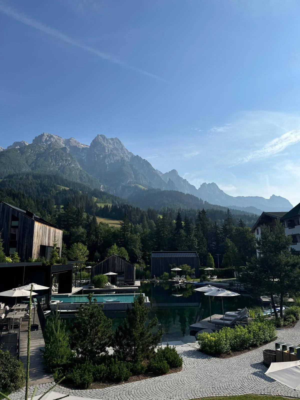 Mountain resort with pool, umbrellas, lounge chairs, wooden buildings, surrounded by trees and forest, with tall mountains in the background under a clear blue sky.