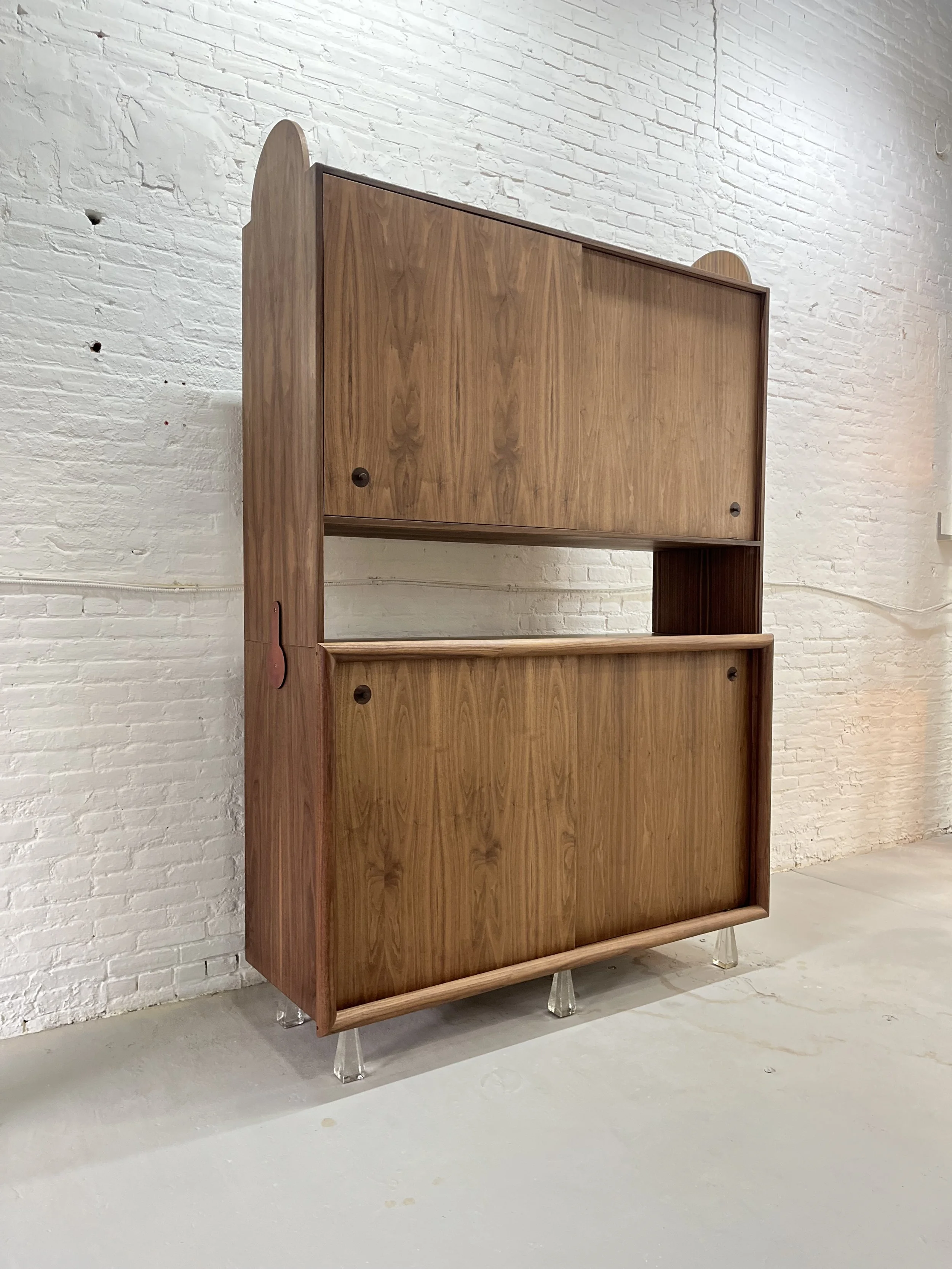 A vintage wooden cabinet with a closed upper compartment and an open lower space, standing on clear, angled legs against a white brick wall.