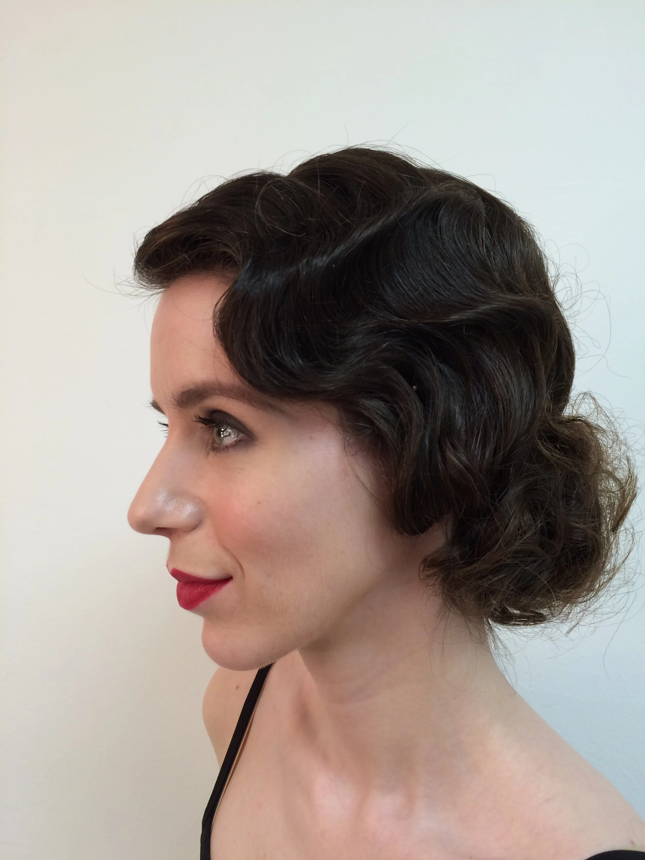Profile of a person with vintage-style short wavy hair, wearing red lipstick, against a plain light background.