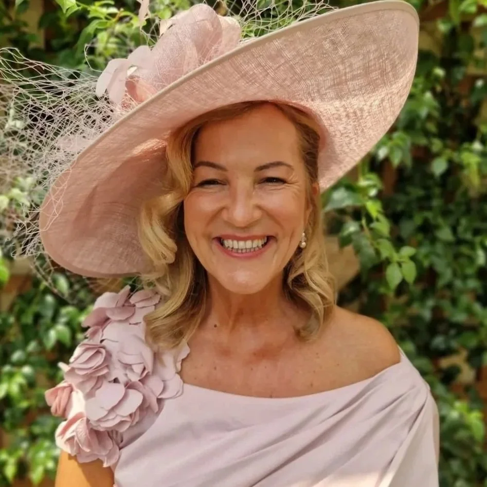 Woman with shoulder-length wavy brown hair, wearing a white floral top, outdoors with trees in the background.