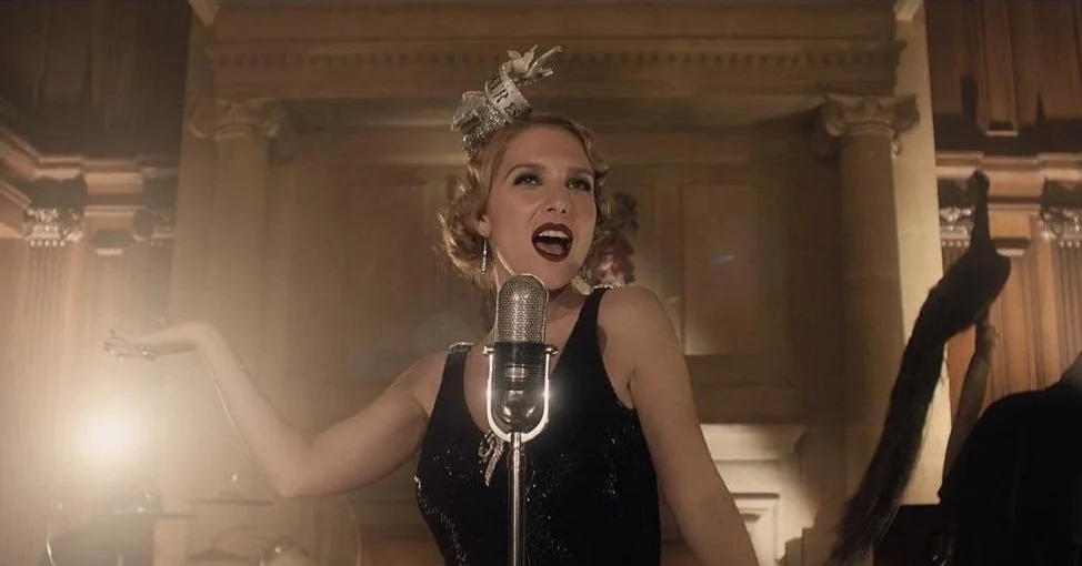 Woman performing in vintage style, wearing a headpiece and black dress, singing into a retro microphone in a dimly lit, elegant setting.