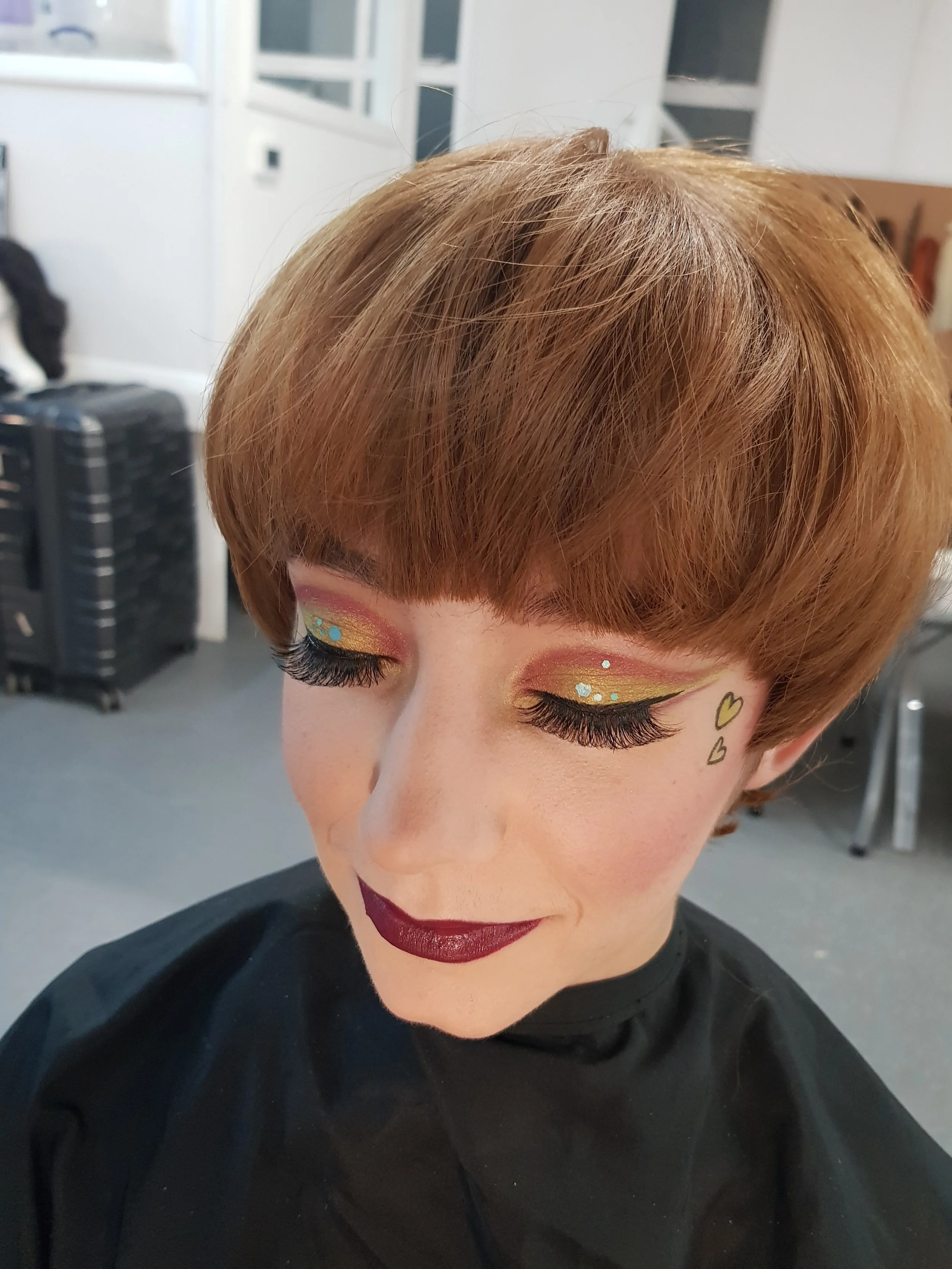 Person with bob haircut and bold makeup, featuring dark lipstick, gold eyeshadow, long false eyelashes, and heart designs on the cheeks.