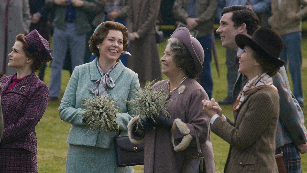 Actors from The Crown, including The Queen and The Queen's mother in vintage clothing, smiling and standing outdoors, holding greenery bouquets.
