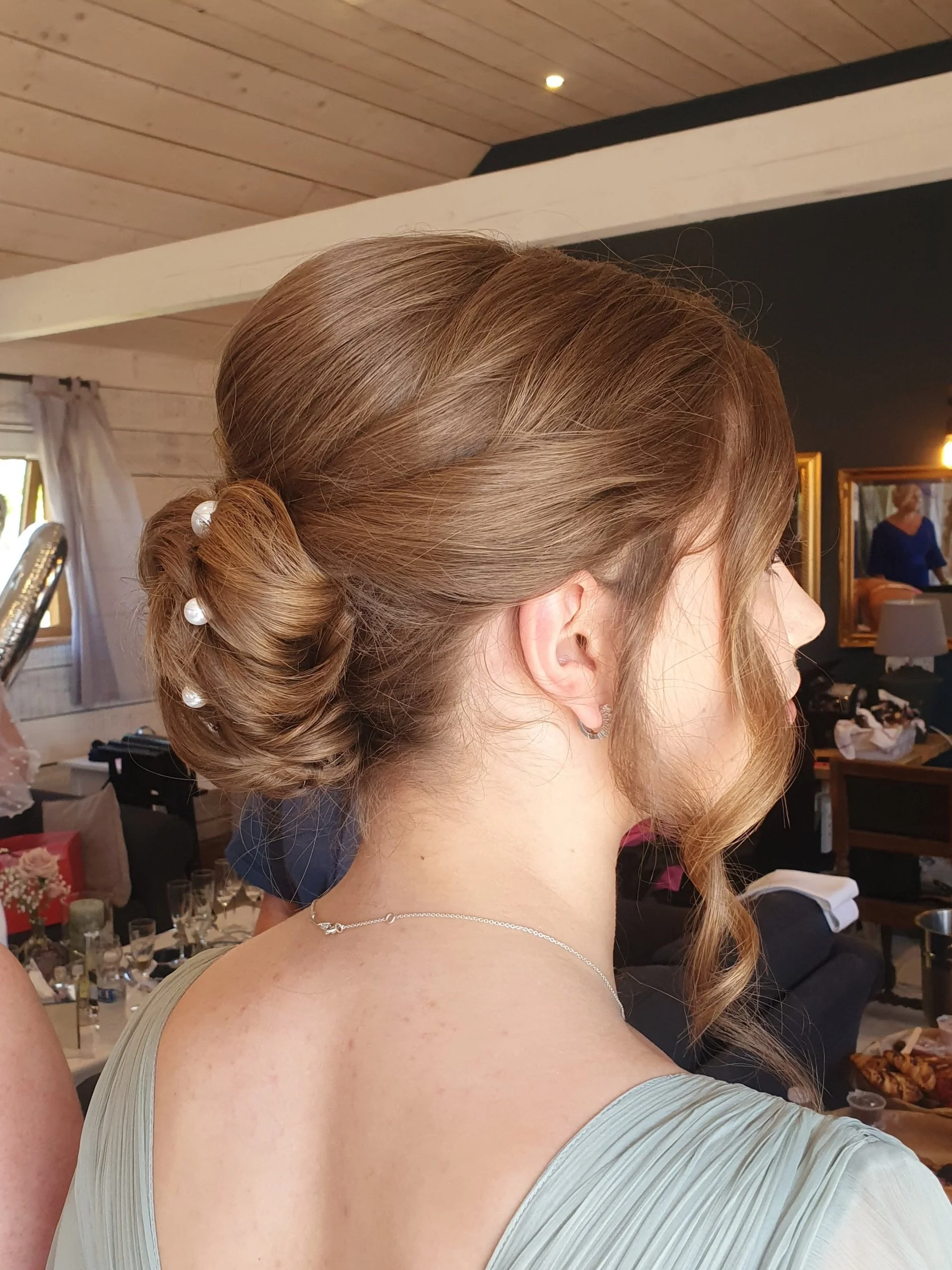 Woman with elegant updo hairstyle adorned with pearls, wearing a silver necklace, in an indoor setting.