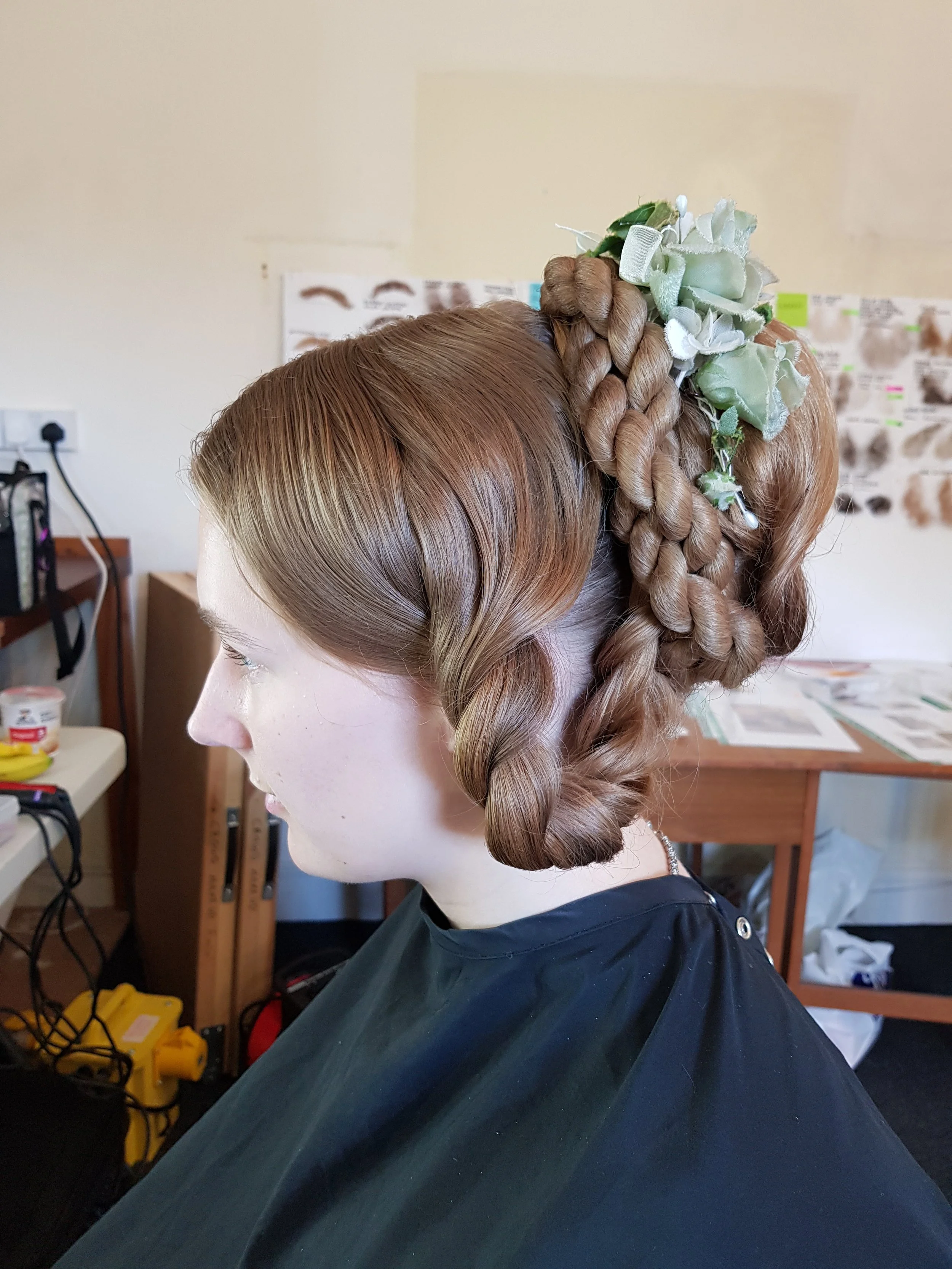 Side view of a person with an intricate braided hairstyle adorned with green foliage, seated in a salon environment.