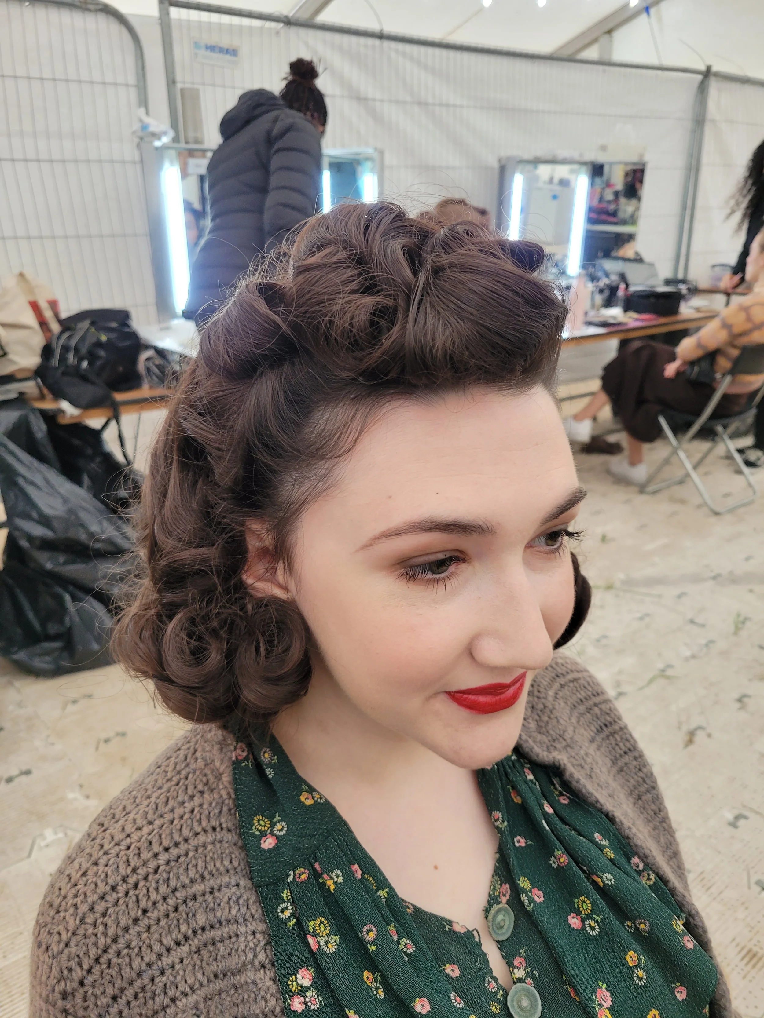 A woman with vintage-style curly hair and red lipstick wears a green floral dress. She sits in a dressing room with people and mirrors in the background.