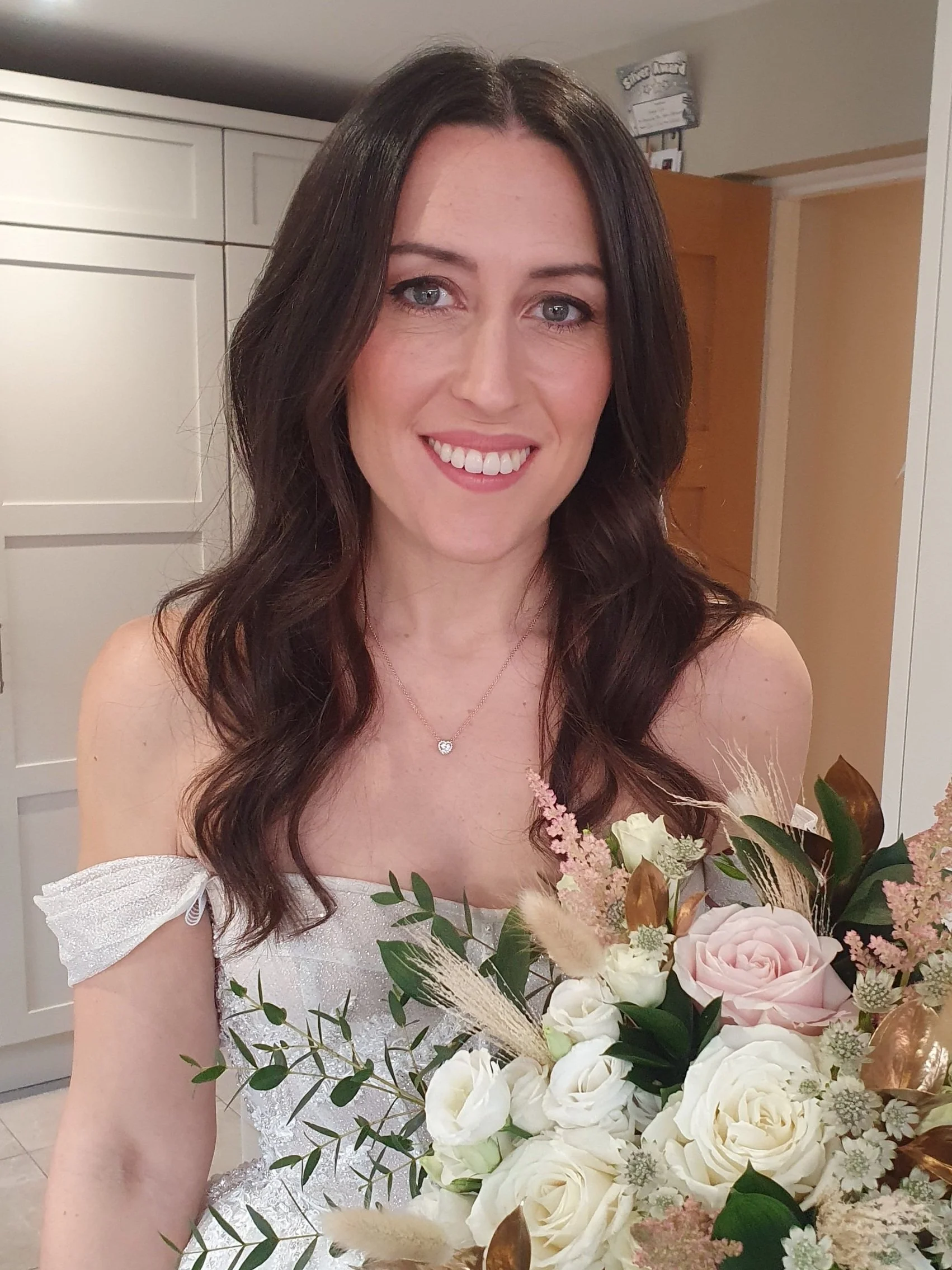 Woman in a white dress holding a bouquet of flowers. The bouquet includes white and pink roses, greenery, and some dried plants. She is smiling, with long dark hair.