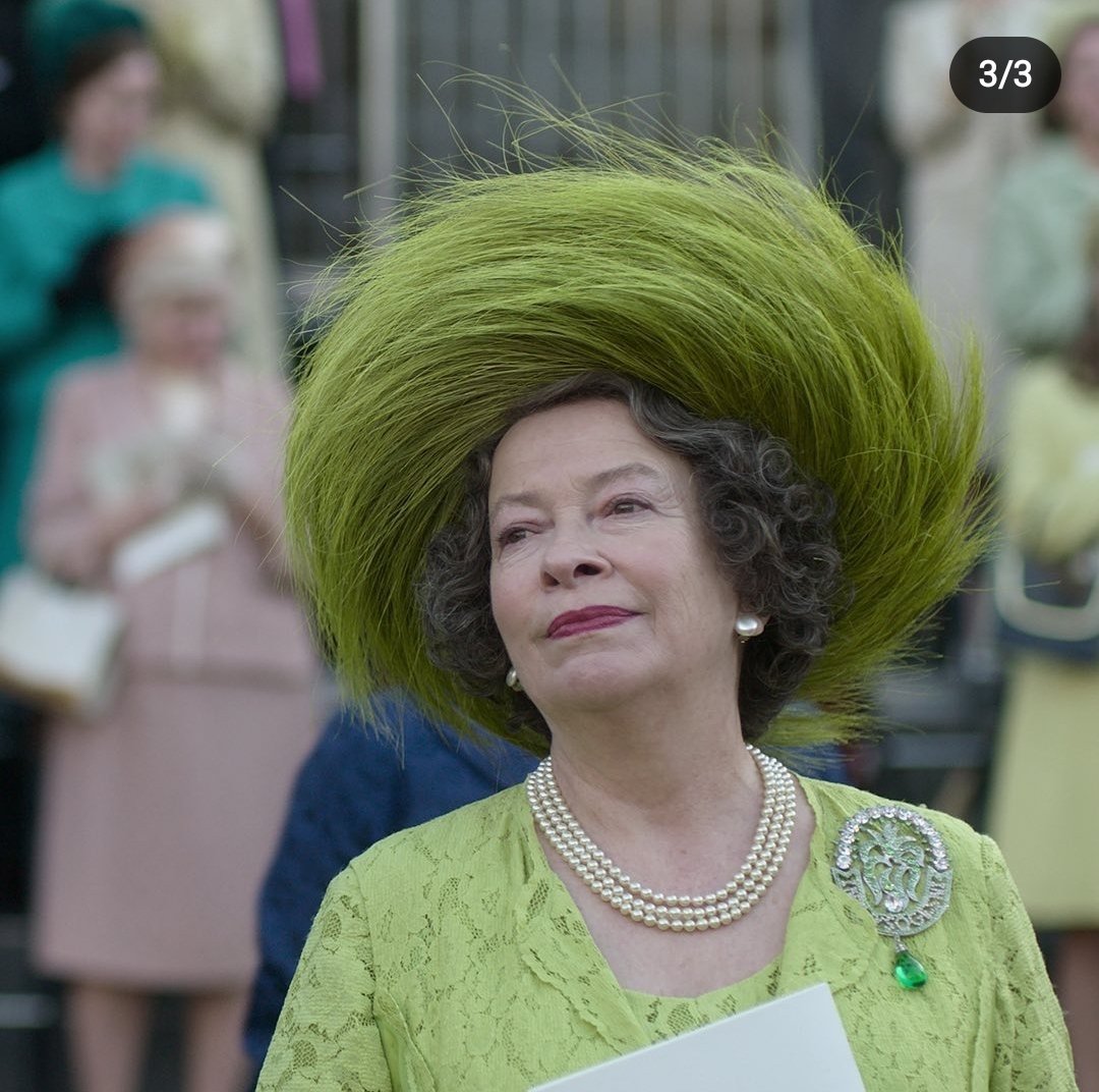 Queen Mother from The Crown wearing an elaborate green hat and matching dress with pearl necklace and decorative brooch.