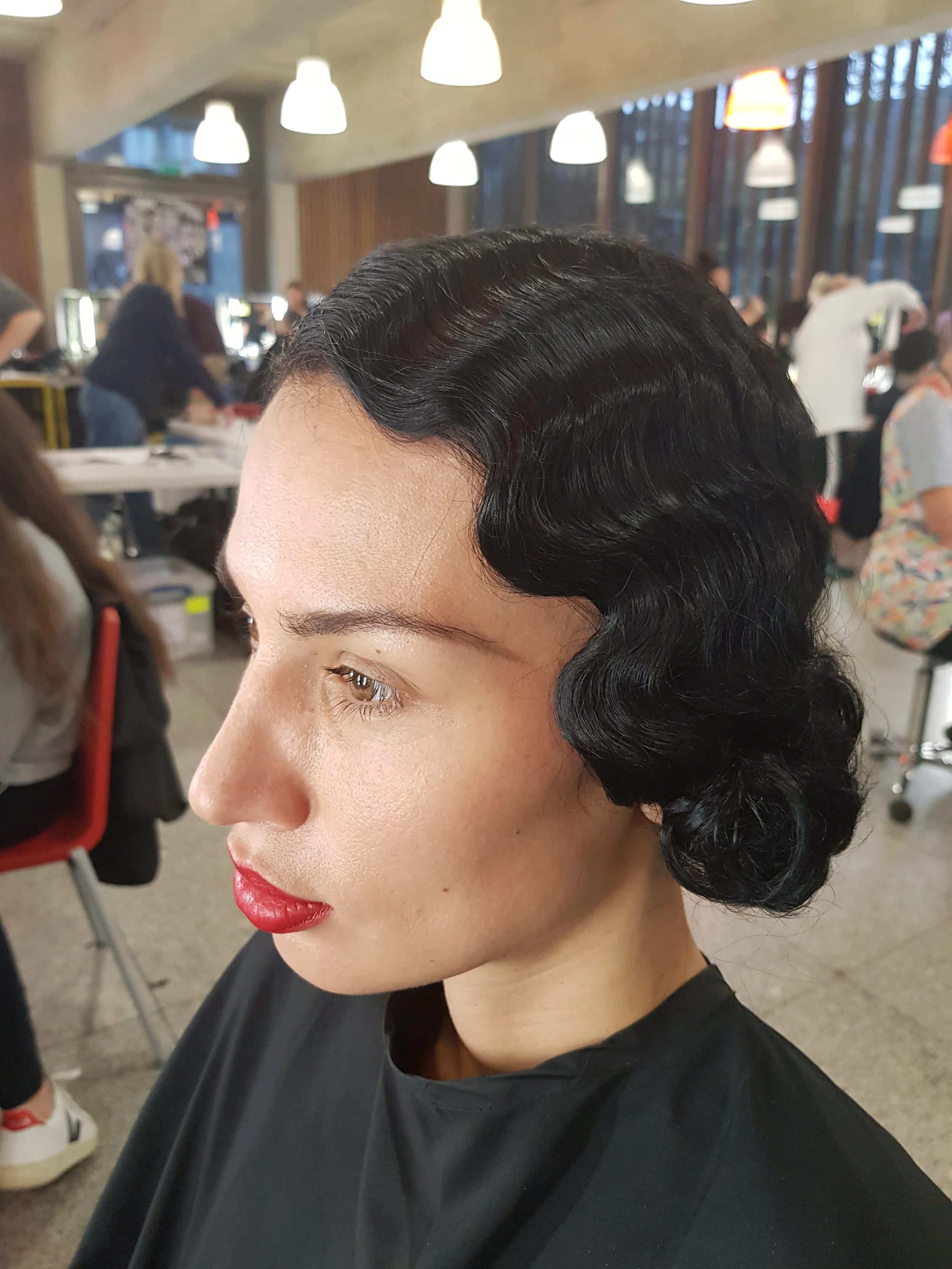 A close-up of a woman with vintage finger wave hairstyle, red lipstick, and a black cape in a salon setting.