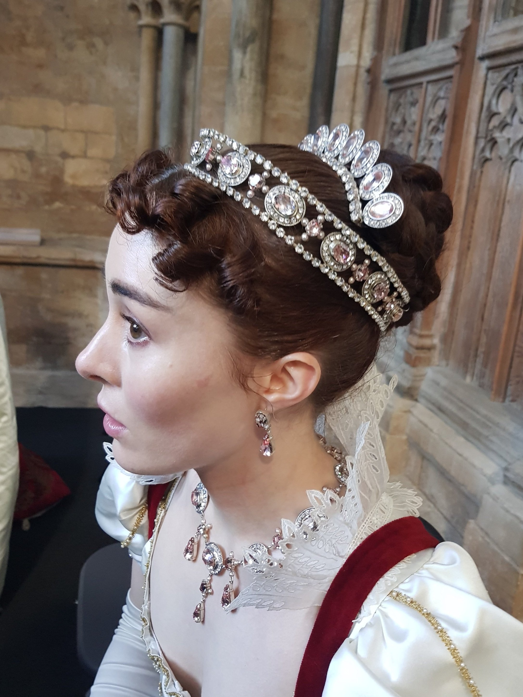 Woman in historical costume with jeweled tiara and necklace, wearing white and red dress, ornate architectural background.
