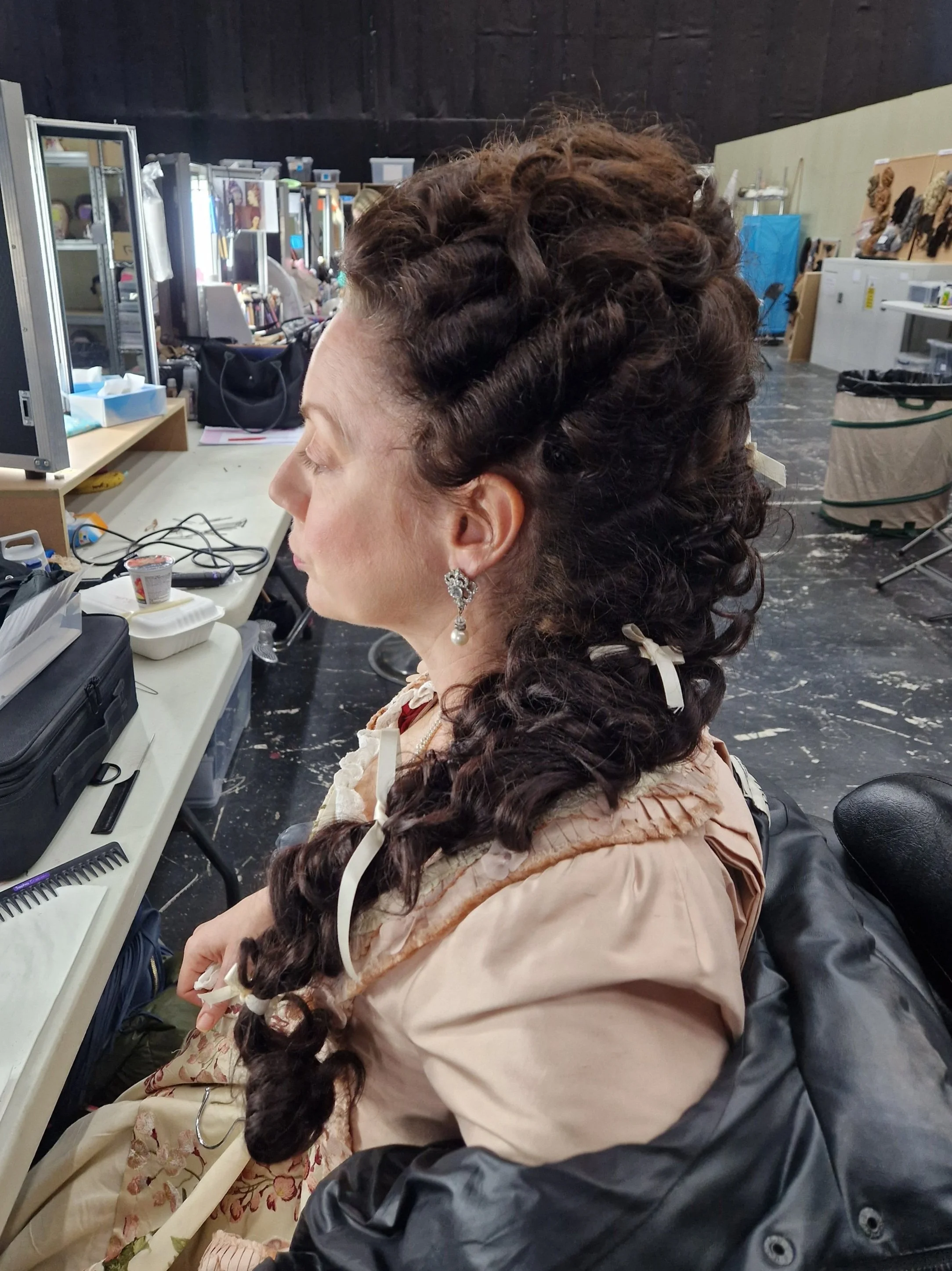 Woman in historical costume with elaborate braided hairstyle and ribbons, sitting in a dressing room.