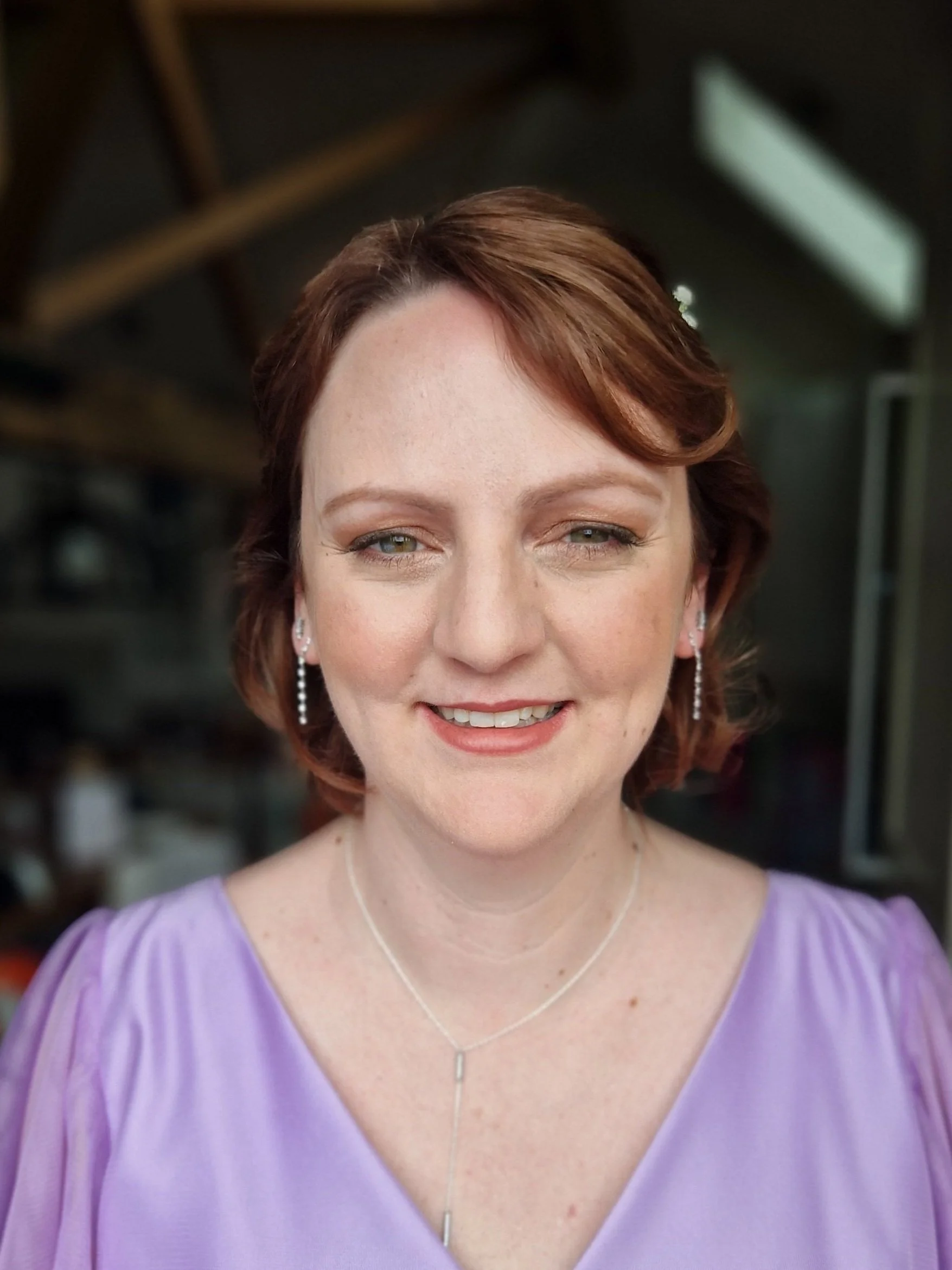 Woman smiling, wearing a light purple dress and delicate jewelry, indoors.