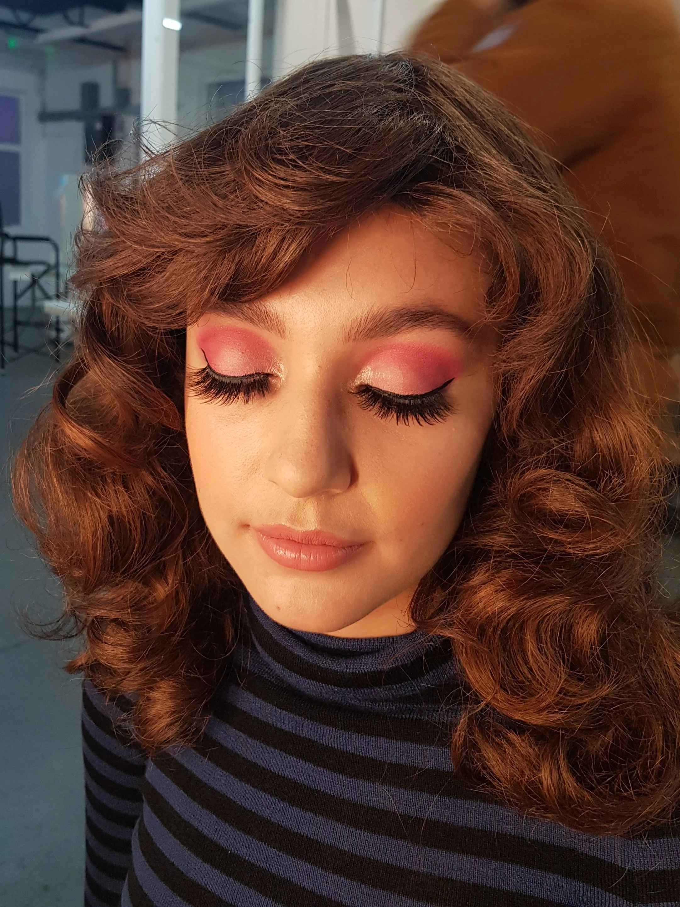 Person with curly hair and pink eyeshadow, wearing a black and blue striped top, eyes closed.