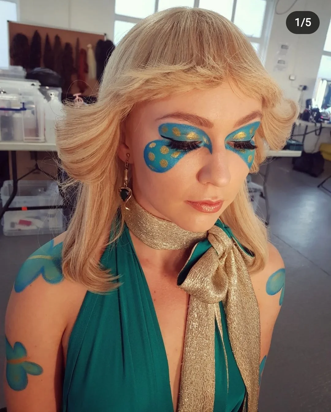 Woman with dramatic blue and gold face makeup, wearing a teal dress and gold scarf, in a room with various items in the background.