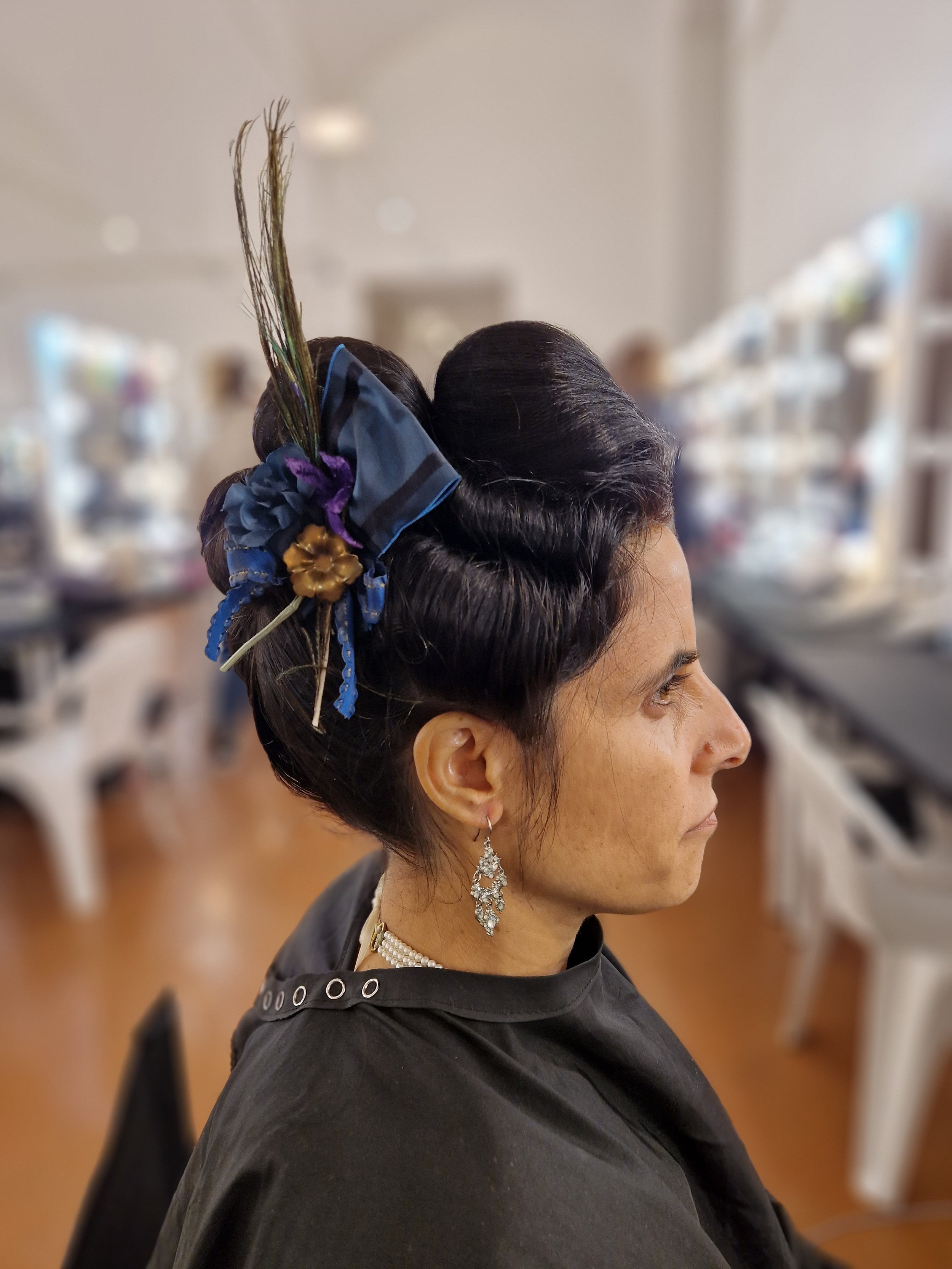 Woman with an elaborate updo hairstyle featuring a peacock feather and decorative accessories, wearing chandelier earrings and a black salon cape, inside a salon.