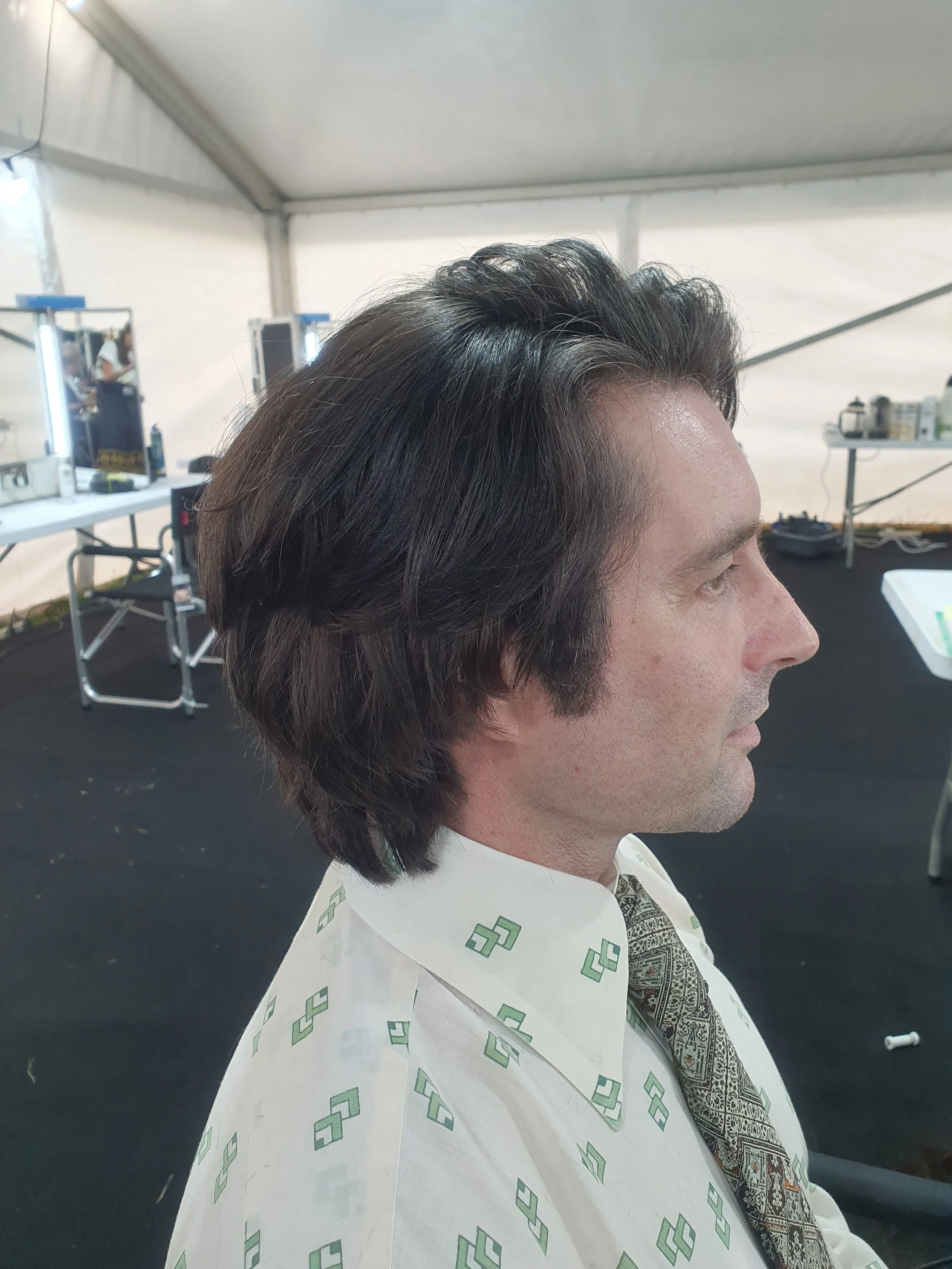 Side profile of a person with dark hair in a tent, wearing a patterned shirt and tie, sitting in front of a mirror and salon equipment.