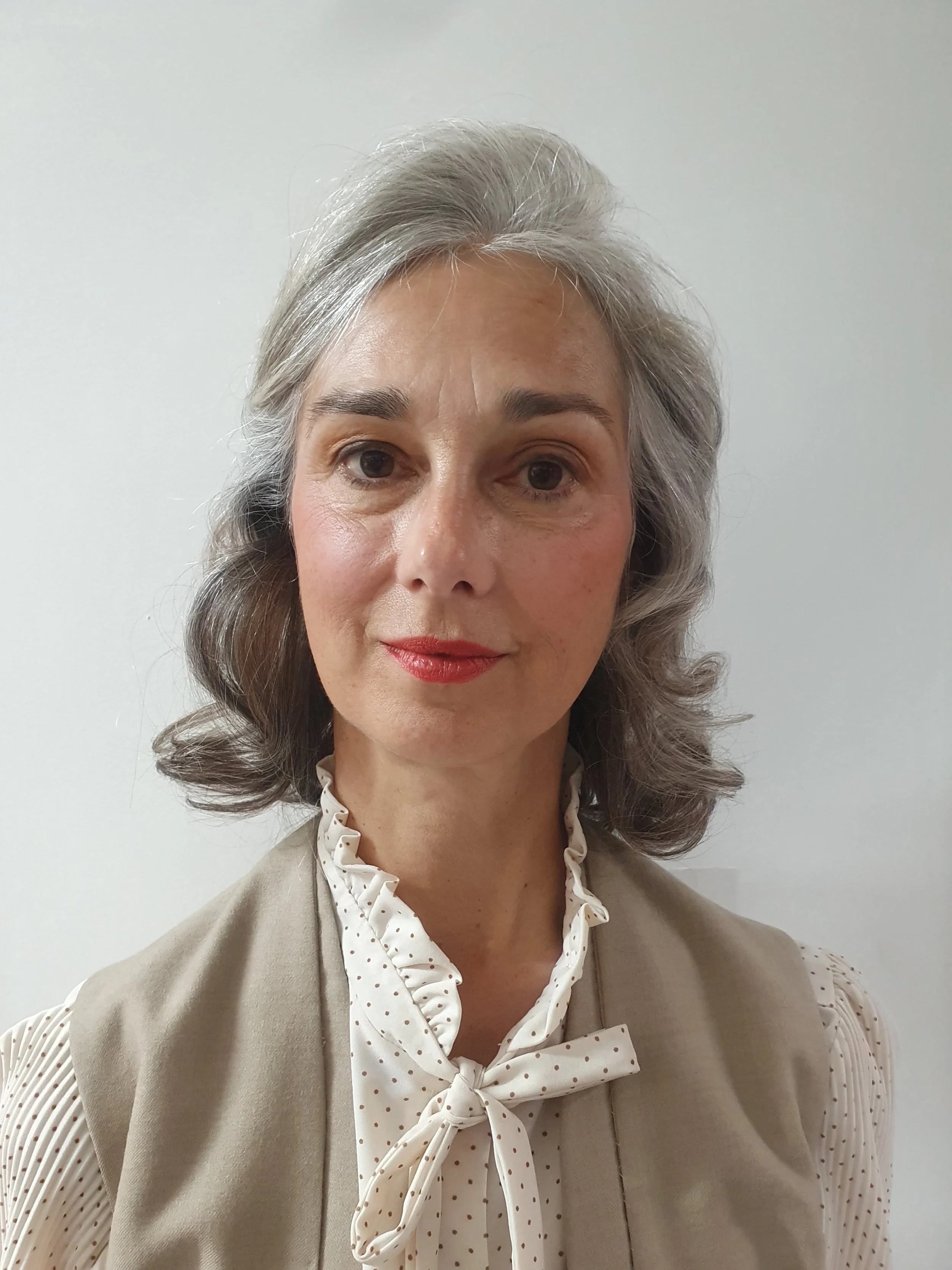 Woman with gray hair, wearing a polka-dot blouse and beige vest, standing against a plain background.