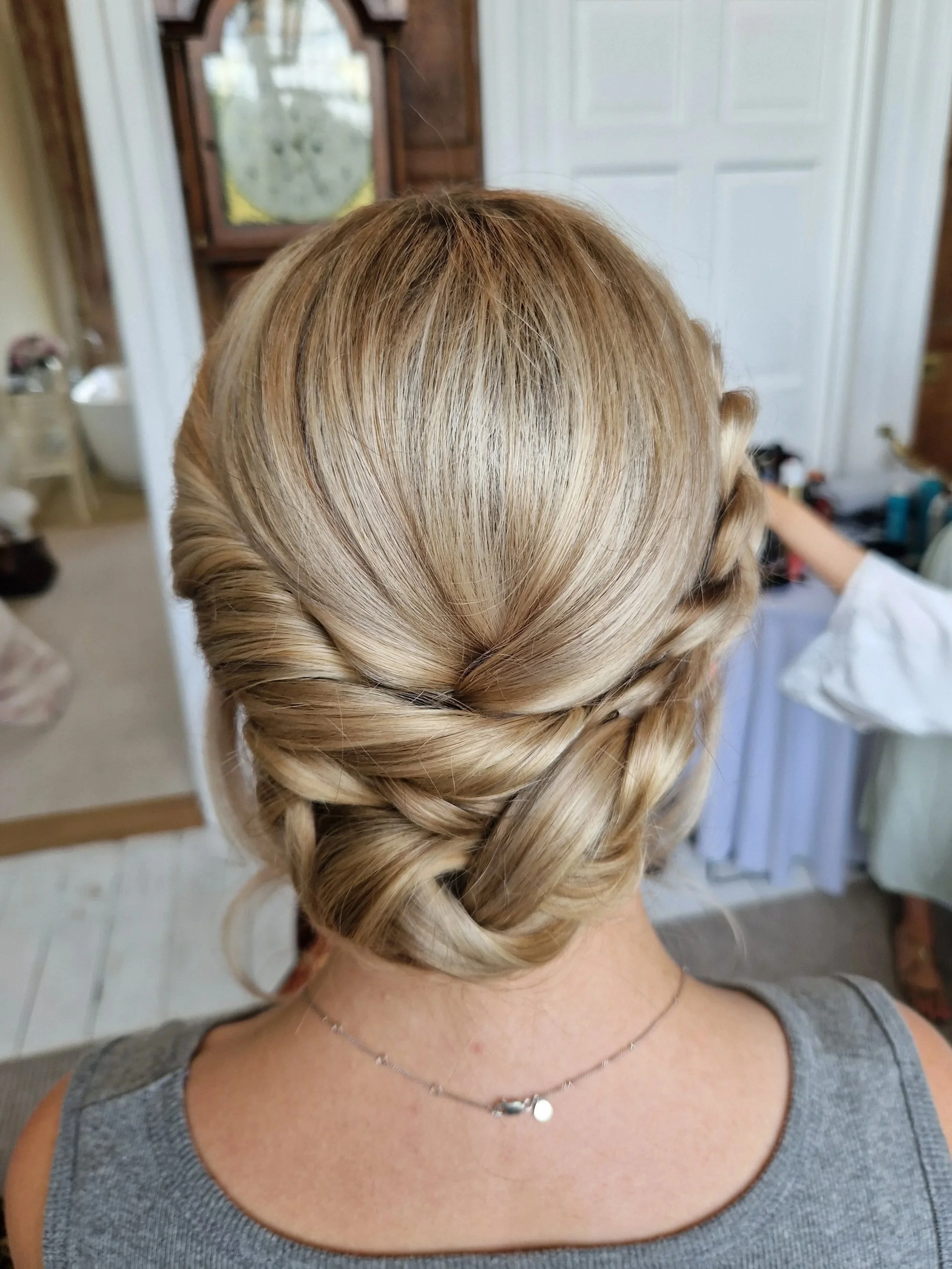 Back view of a woman with an intricate braided updo hairstyle, wearing a gray top and a delicate necklace, in a room with a mirror and wooden paneling.