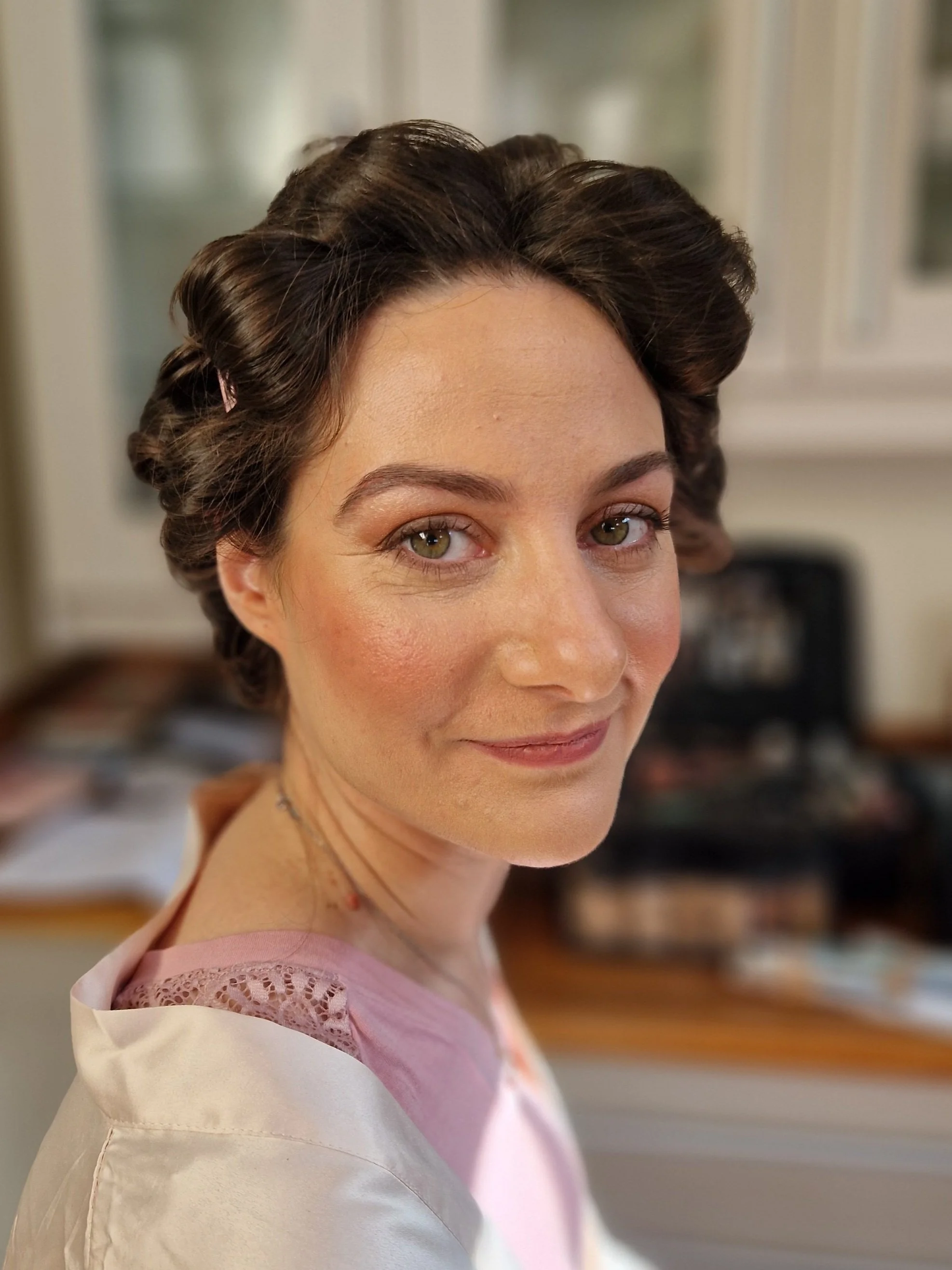 Person with styled hair, softly smiling, wearing a white garment over a pink dress, indoors with makeup case in background.