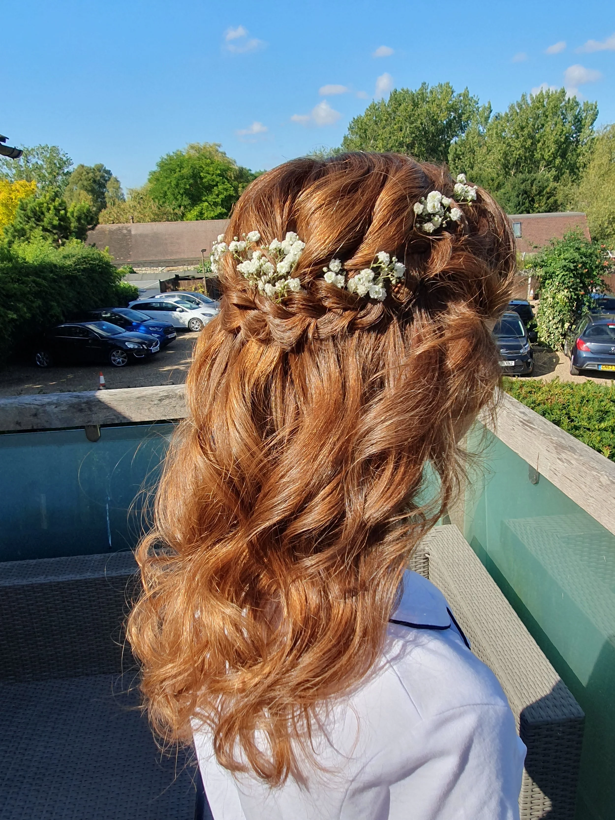 Back view of a person with red hair styled in loose curls and a braided crown adorned with small white flowers, outdoors on a sunny day.