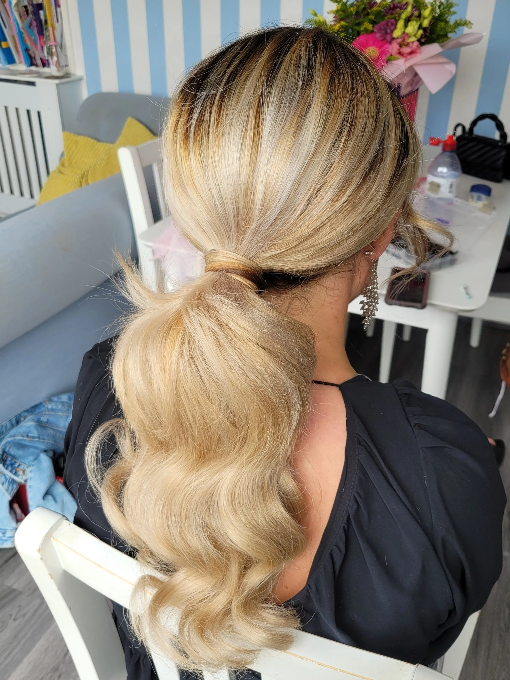 Woman with blonde hair in a ponytail, seated indoors, with flowers and items on the table.