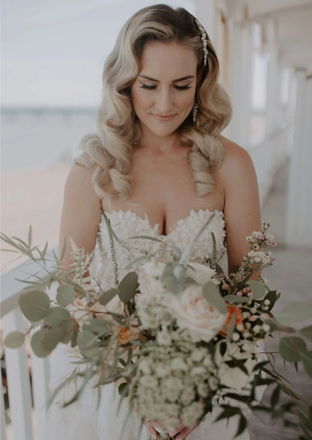 Bride holding a large bouquet of flowers, dressed in a white lace wedding gown, with long wavy blonde hair and a decorative hairpin, on a balcony.