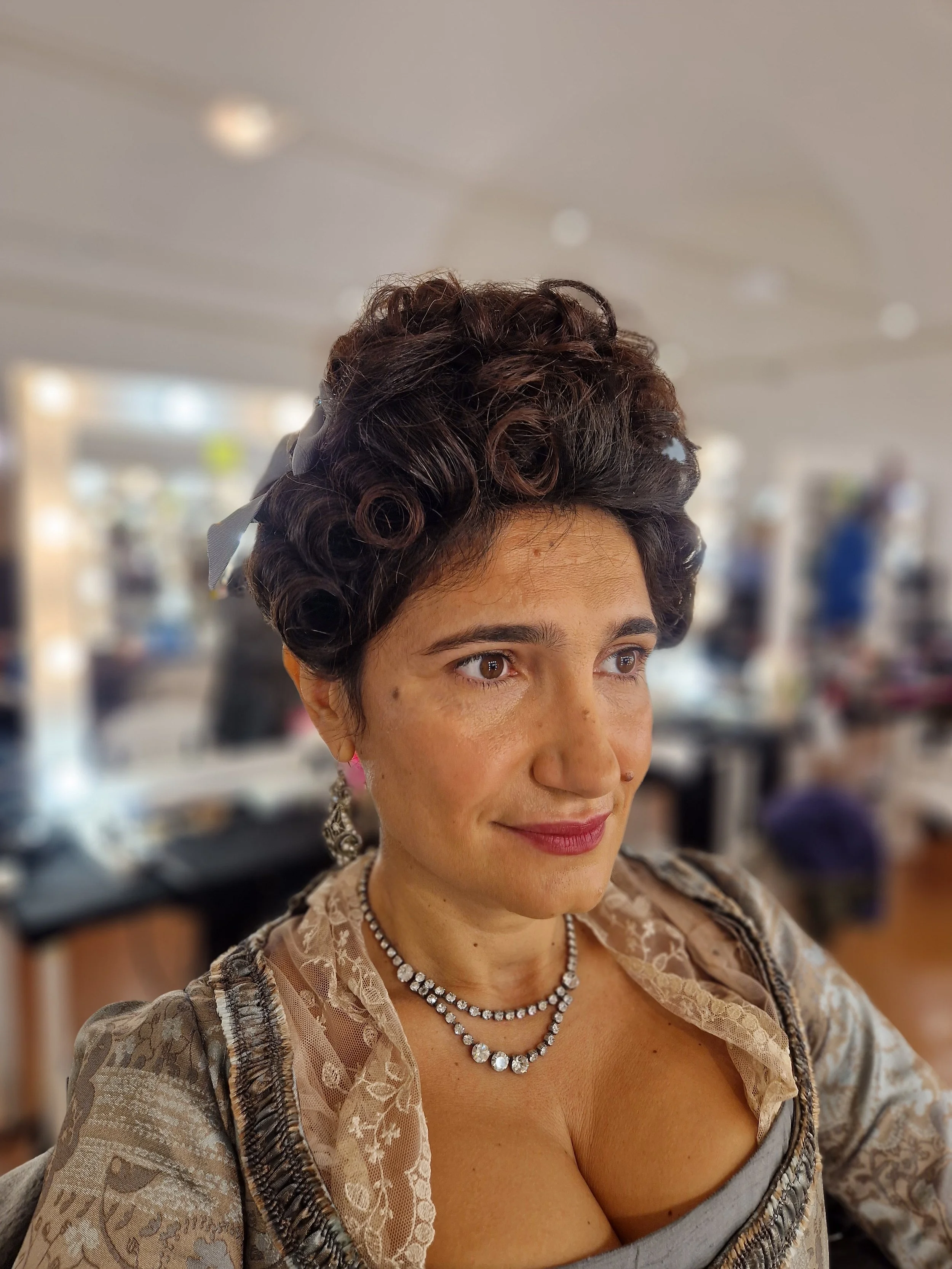 Person in historical costume with curly hair, lace dress, and pearl necklace.