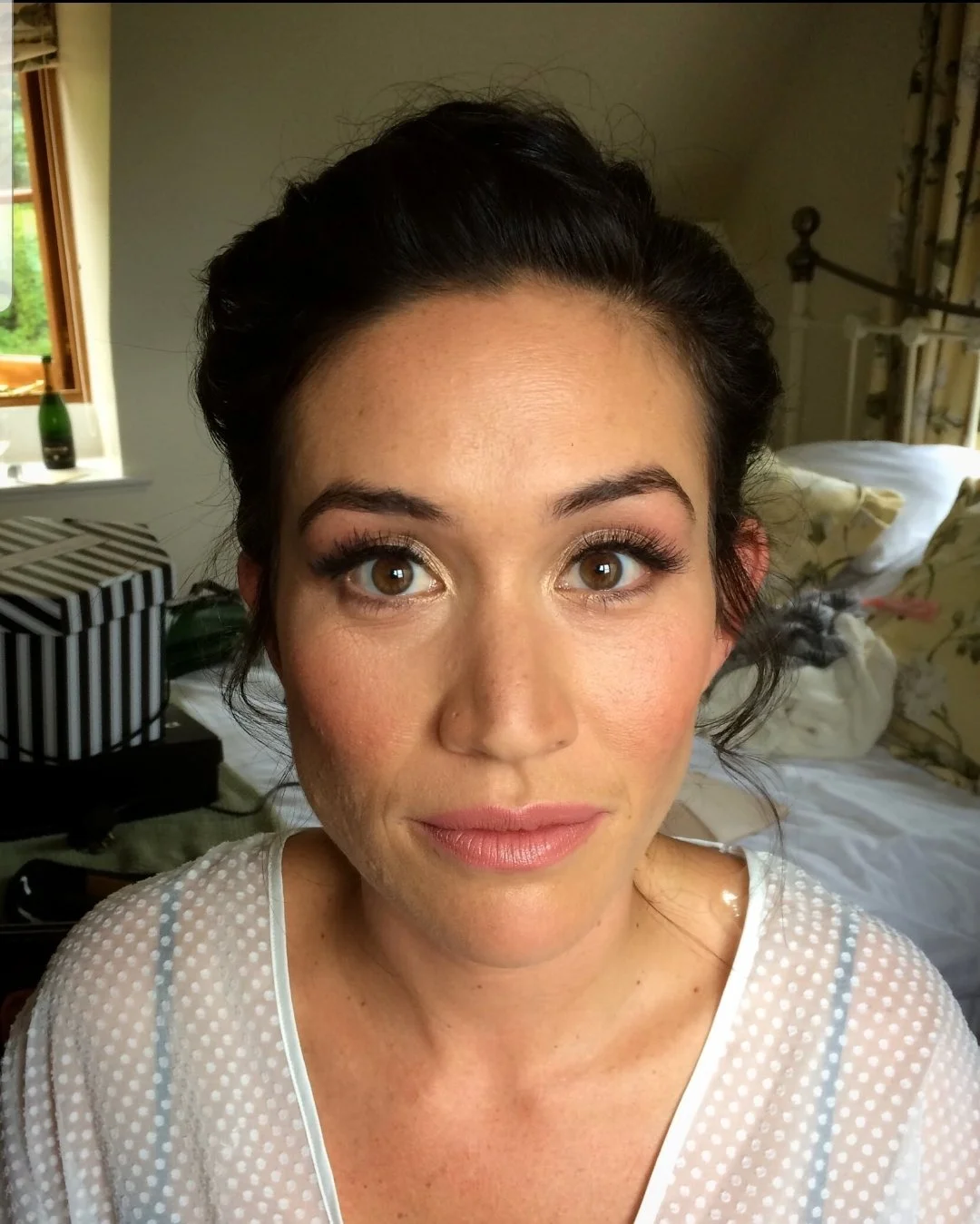 Woman with dark hair styled in an updo, wearing light makeup and a white dotted top, sitting indoors with a window and bed in the background.