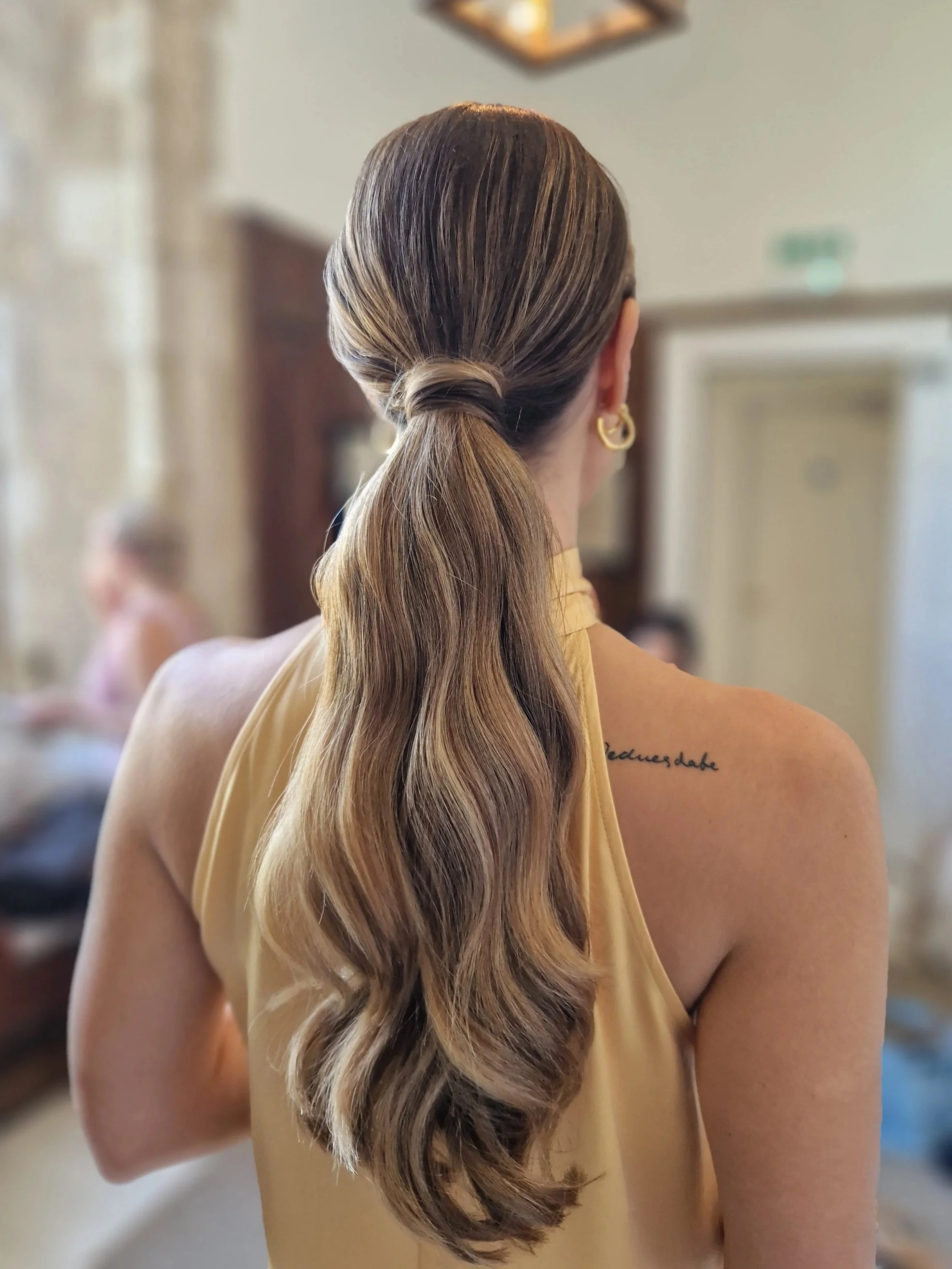 A woman with long, wavy hair tied in a low ponytail, wearing a sleeveless top. The focus is on her hair and back, with a blurred indoor background.