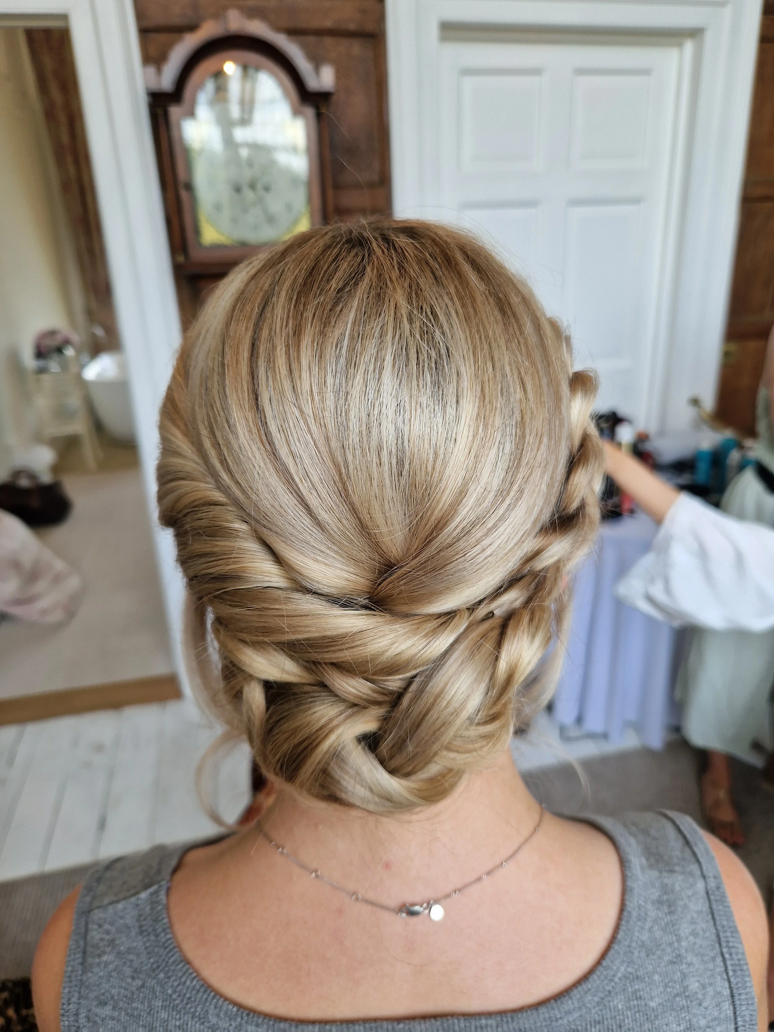 Back view of a woman with an intricate braided updo hairstyle, wearing a gray top and a delicate necklace, in a room with a mirror and wooden paneling.