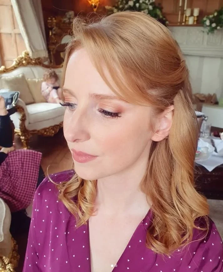 A woman with shoulder-length blonde hair styled in loose curls is wearing a purple top with white polka dots. She appears to be indoors in an elegantly furnished room, possibly with chandeliers and floral decorations.