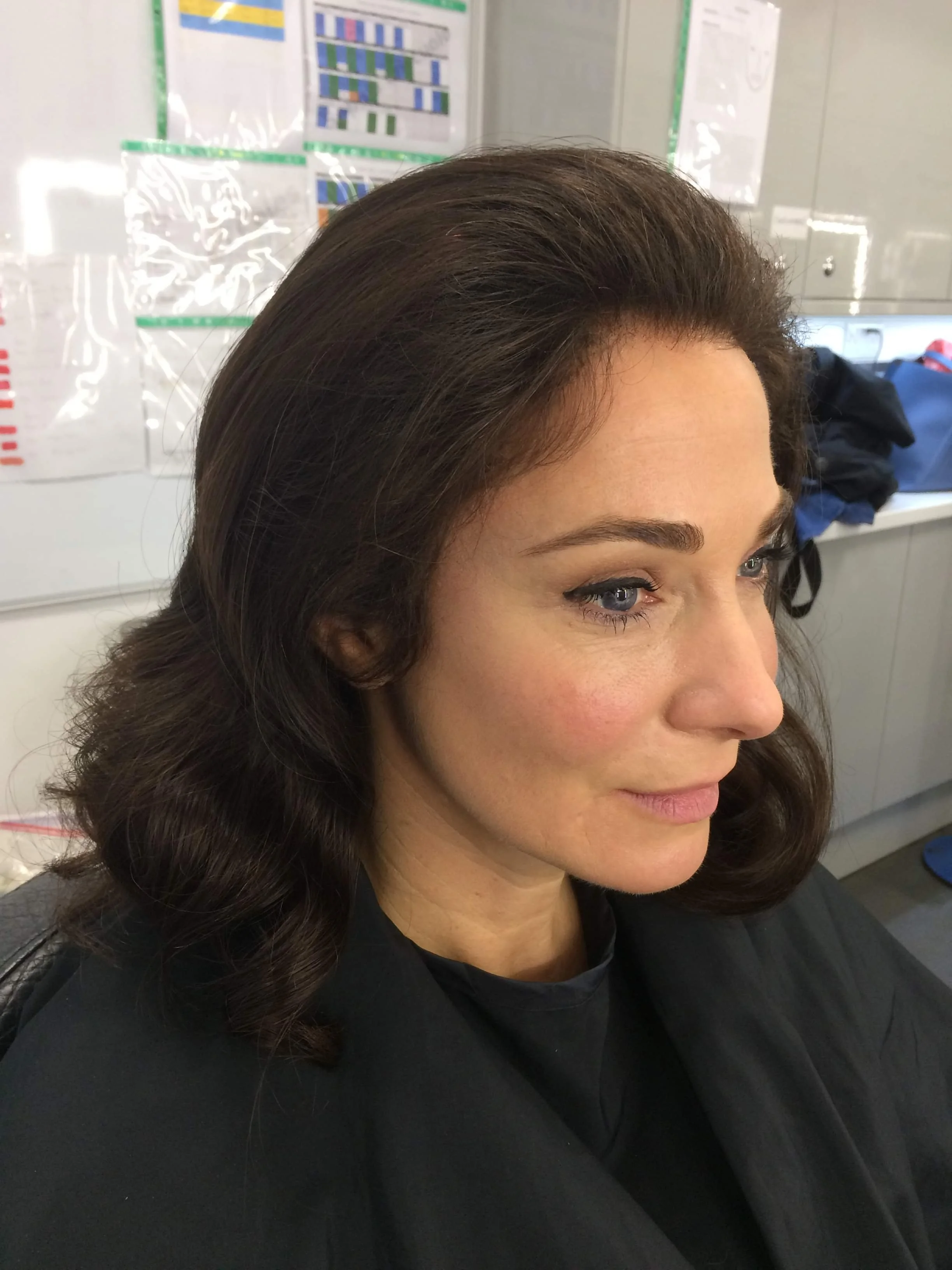 Woman with styled hair wearing a black cape in a room with charts and papers on the wall.
