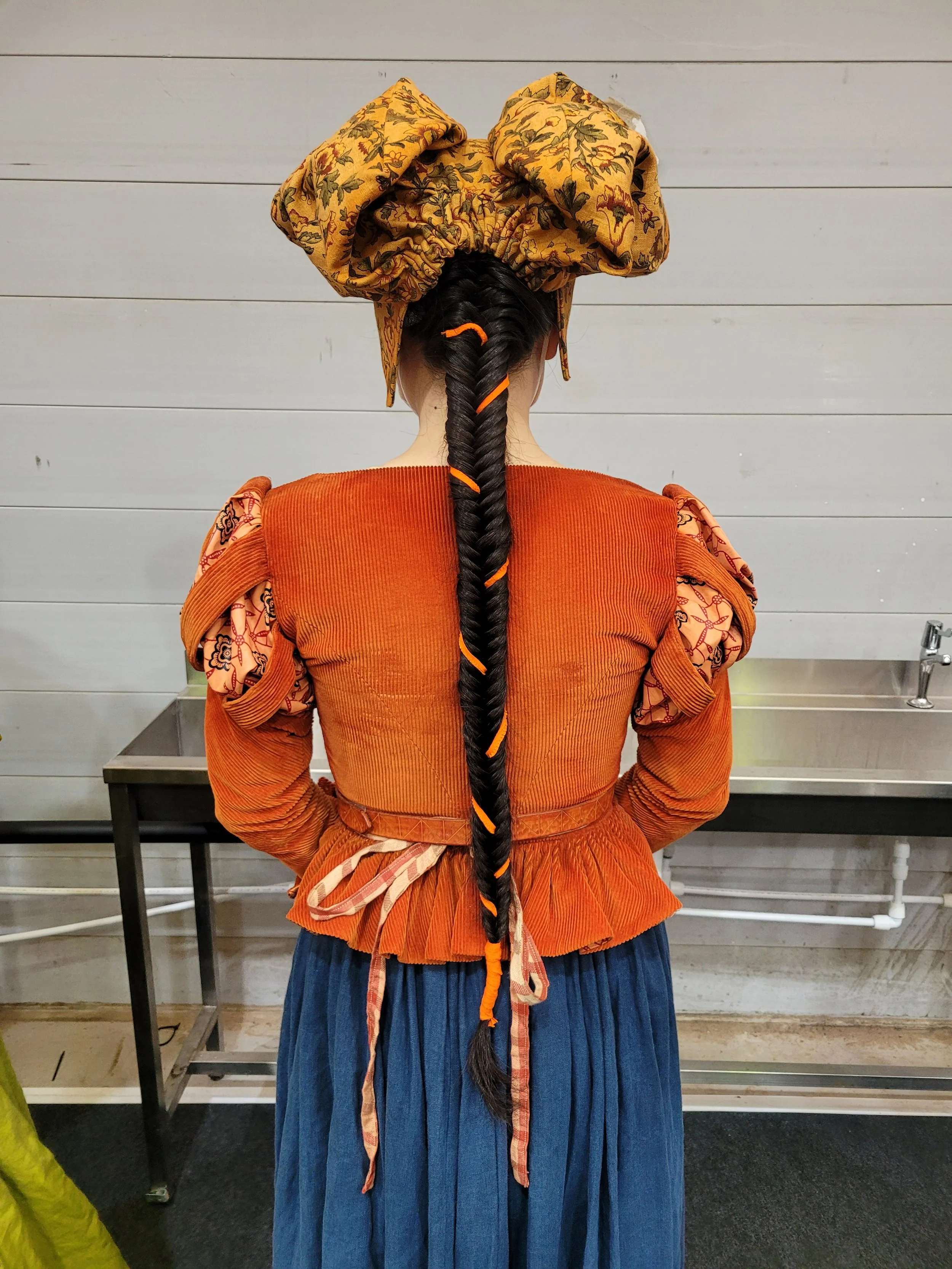 Person with long braided hair and ribbon, wearing a patterned headdress, vintage-style orange top with puffy sleeves, and blue skirt, standing in front of a metal table.
