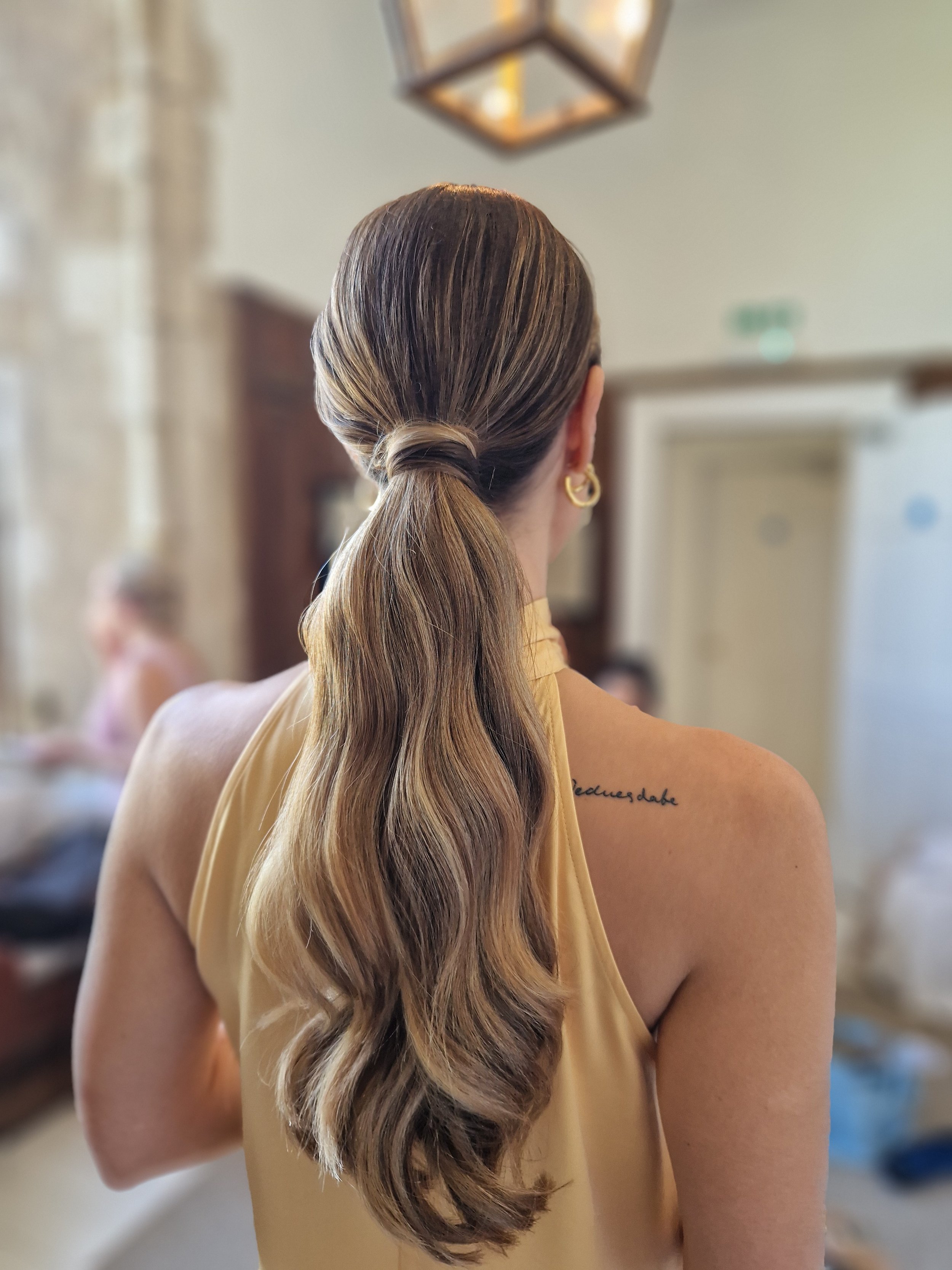 A woman with long, wavy hair tied in a low ponytail, wearing a sleeveless top. The focus is on her hair and back, with a blurred indoor background.