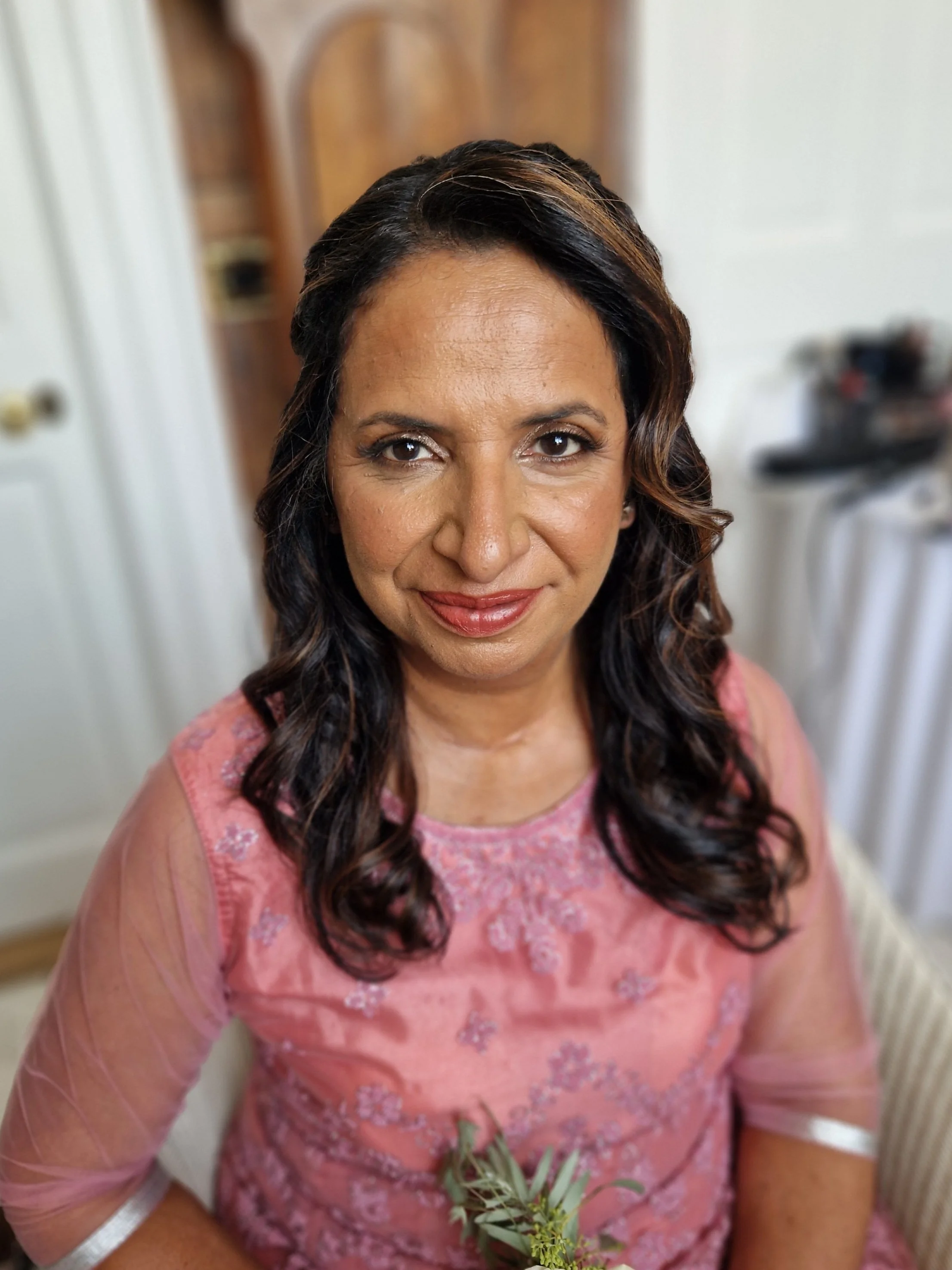 Woman with curly brown hair wearing a pink embroidered dress, sitting indoors, smiling.