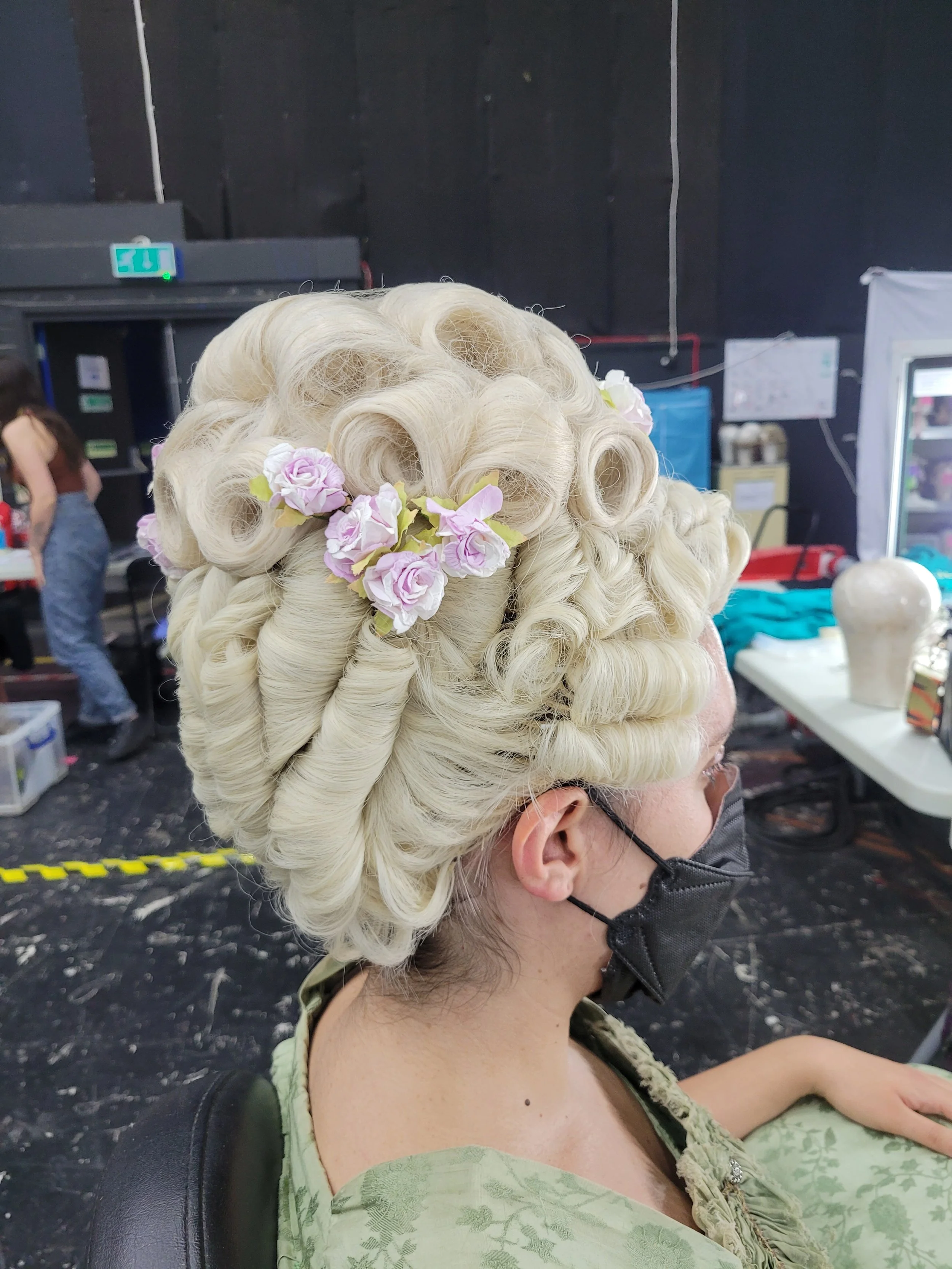 Person wearing an elaborate white wig with curls and pink flowers, sitting in a chair, wearing a face mask and a green patterned dress, inside a backstage area.